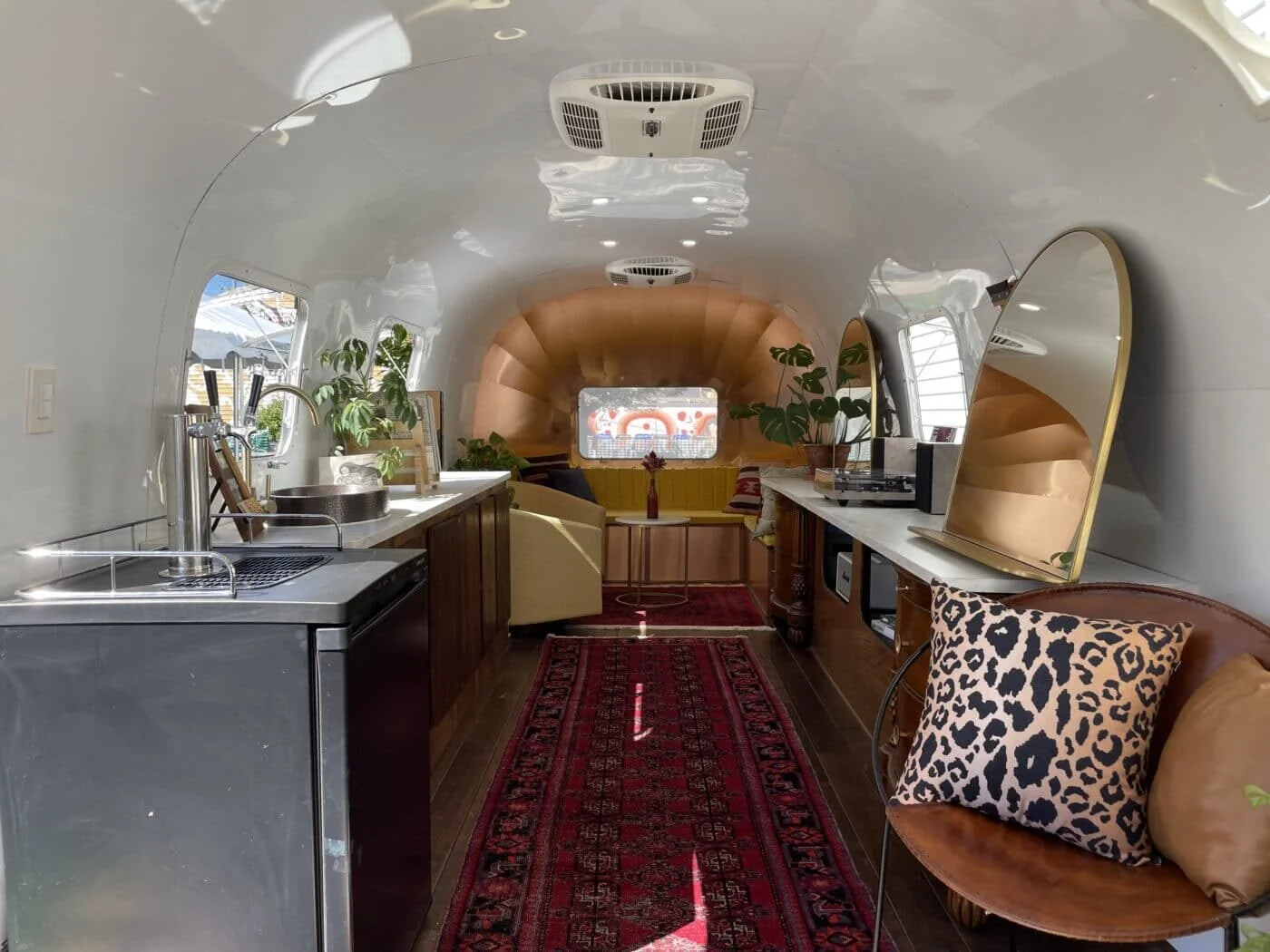Inside view of a tiny home with a rounded ceiling, featuring a kitchen area with a sink, potted plants, and a long countertop on the right, and a cozy seating area at the back with a small window, a yellow bench, and a round table.