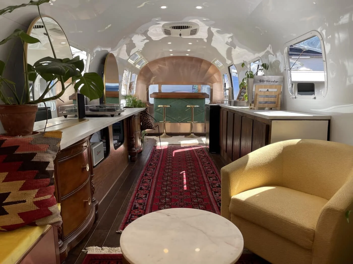 Interior of a vintage Airstream trailer converted into a cozy living space with a yellow armchair, a round marble table, plants, and kitchen area with wood cabinets and a red patterned rug.