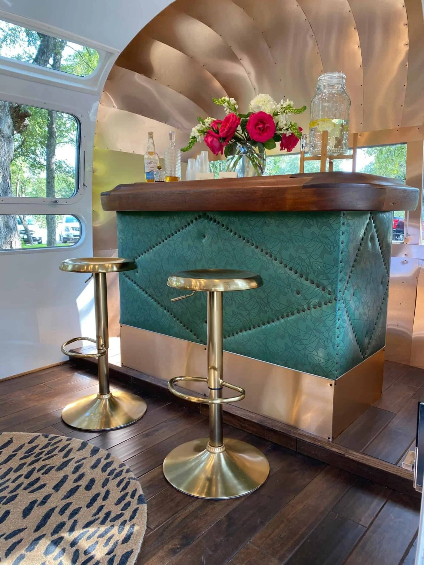 Inside a modern-style mobile home or RV with large windows showing trees outside. There is a bar with a teal upholstered base and a wooden countertop, decorated with a flower vase containing pink and white flowers. Two gold-colored bar stools with ro