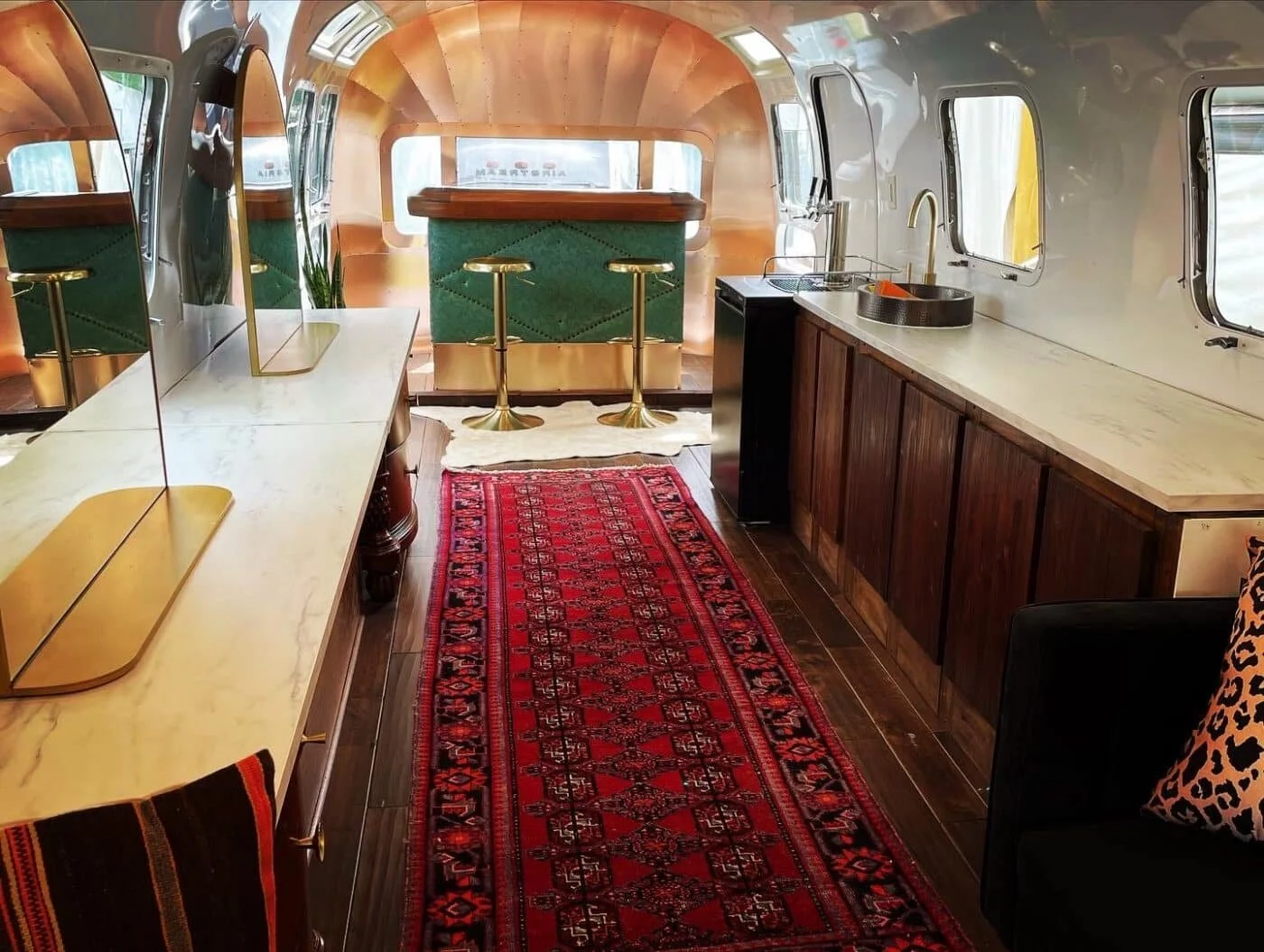 Interior of a converted Airstream trailer with wooden cabinetry, a marble countertop, a sink, a red patterned rug, a black chair with a leopard print pillow, and a small bar area with two green chairs.