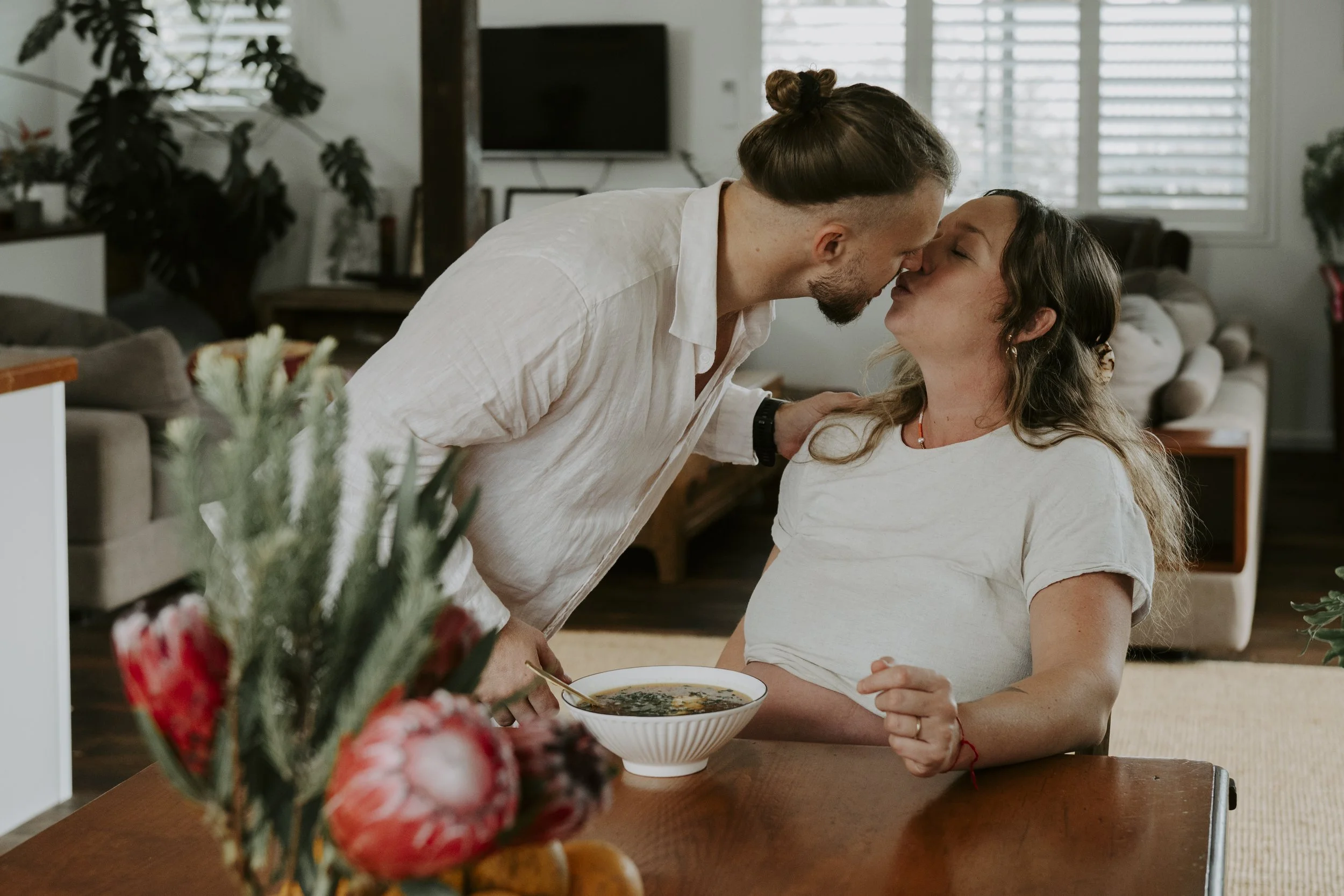 A man leaning over a woman sitting at a dining table, touching foreheads and smiling in a cozy, well-lit living room with large windows and a decorated table with a bowl of soup.