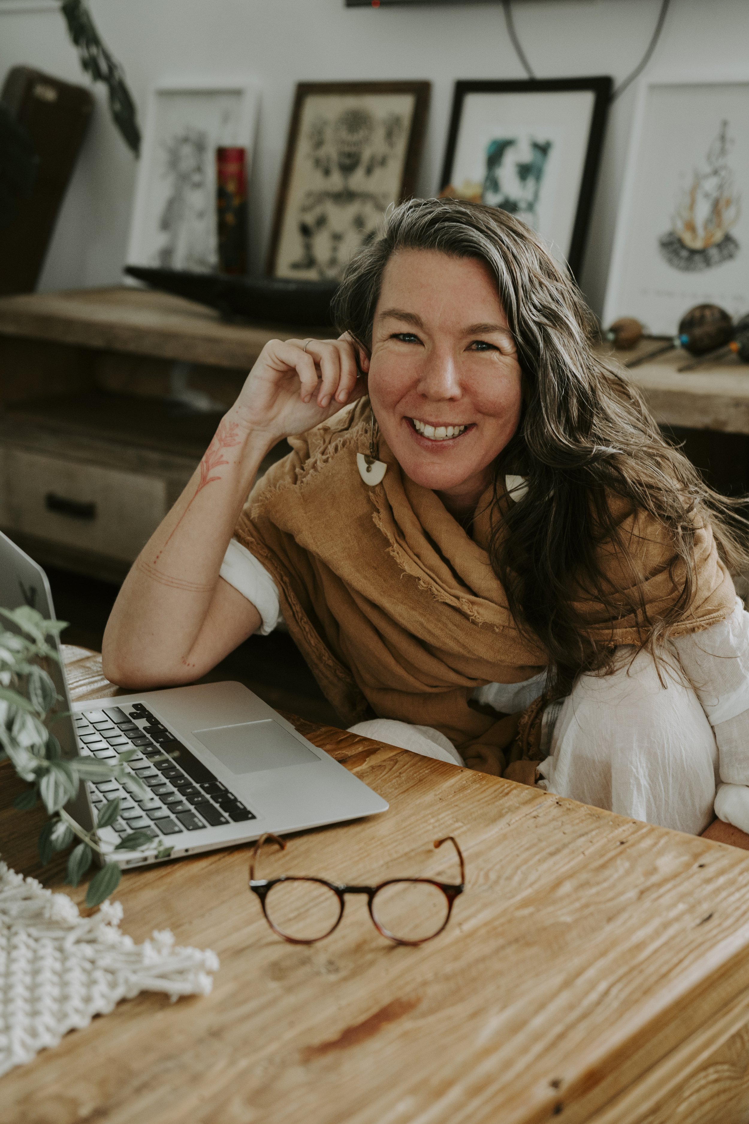 A smiling woman with long, wavy hair, wearing earrings, sitting at a wooden table with a laptop, glasses, and decorative items, in a cozy, art-filled room.
