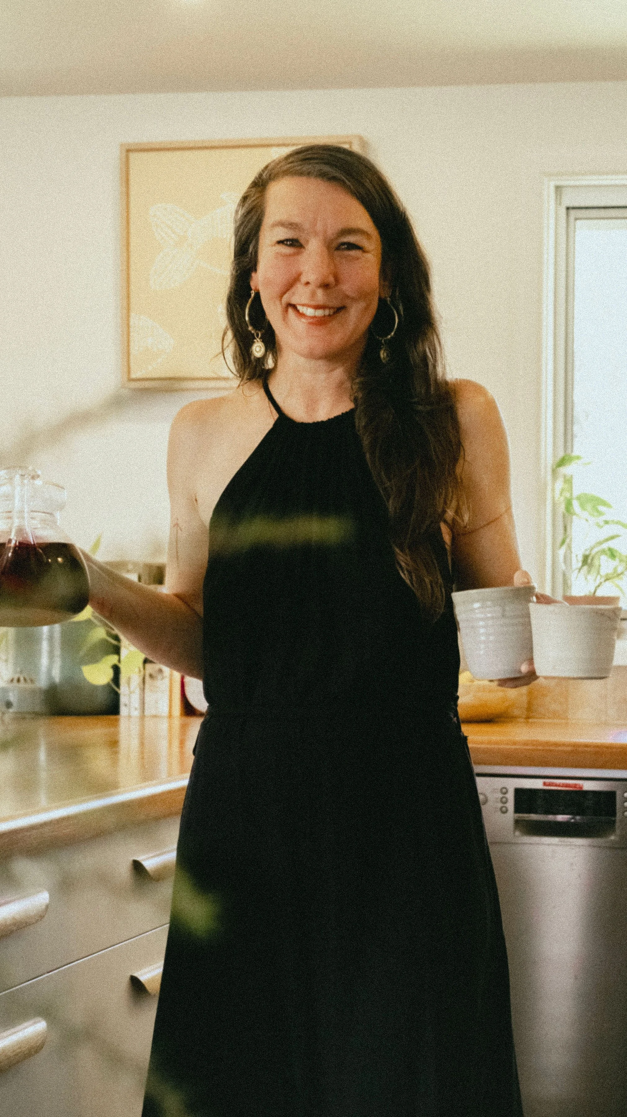 A smiling woman with long brown hair, wearing a black sleeveless dress and hoop earrings, holding a glass carafe of red wine in one hand and a white bowl in the other, standing in a kitchen with white cabinets and a window.