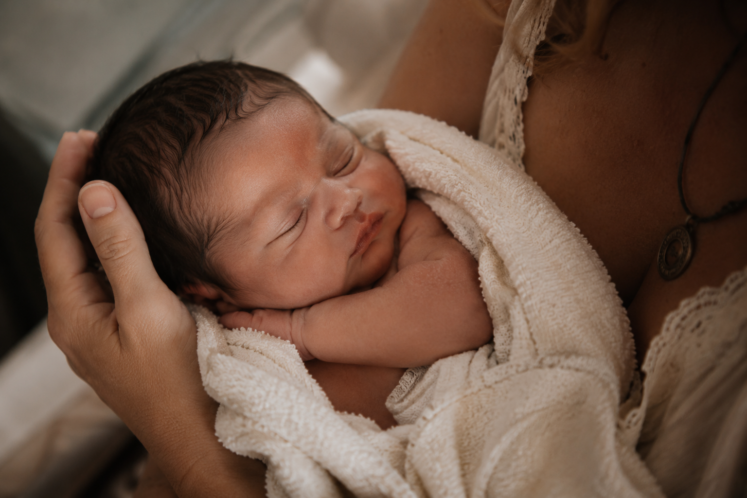 A newborn baby sleeping peacefully, cradled in a person's arms, wrapped in a soft, cream-colored towel.