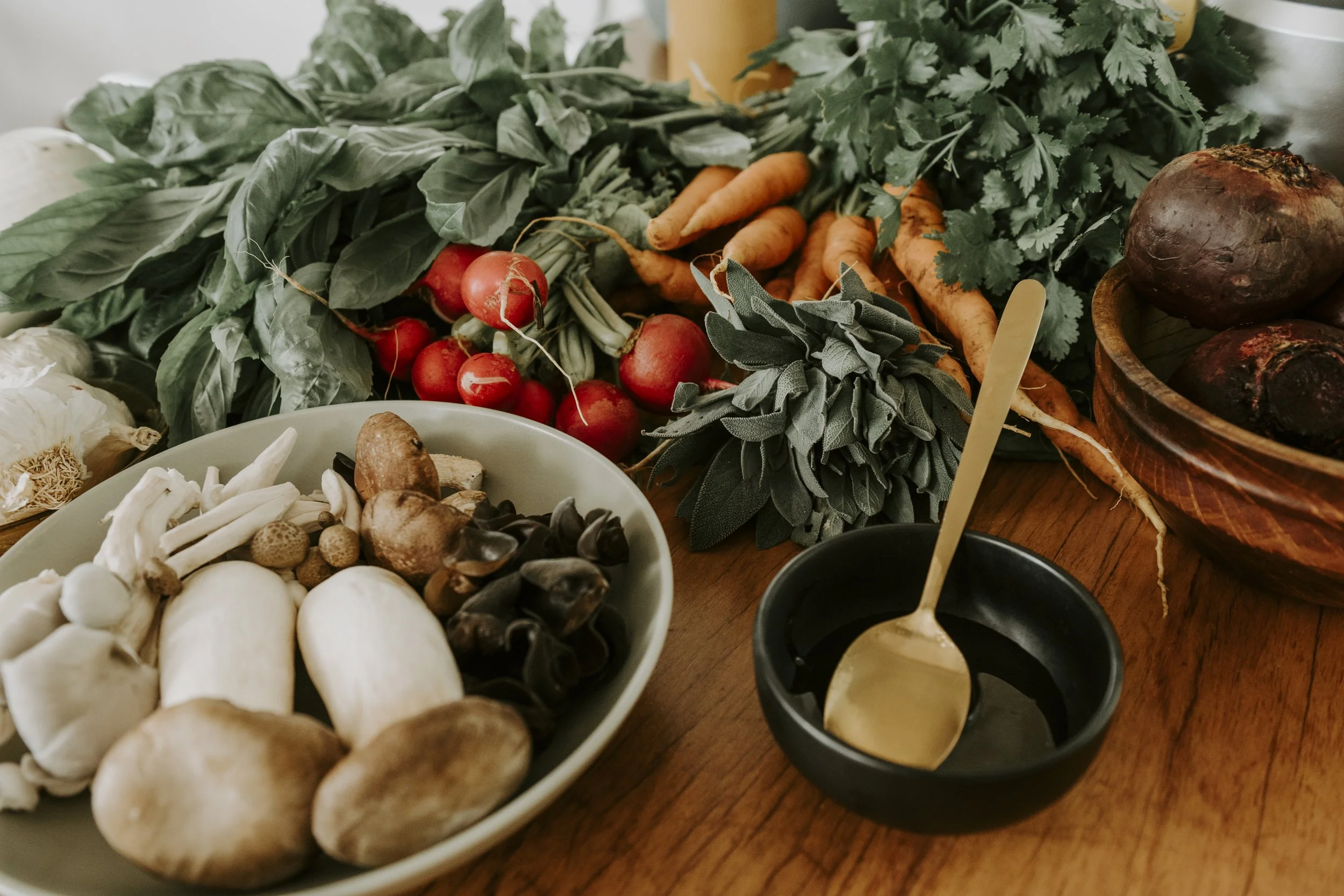 Fresh vegetables on a table including radishes, carrots, spinach, cilantro, and beets, along with a bowl of mushrooms and a small black bowl with dipping sauce and a wooden spoon.