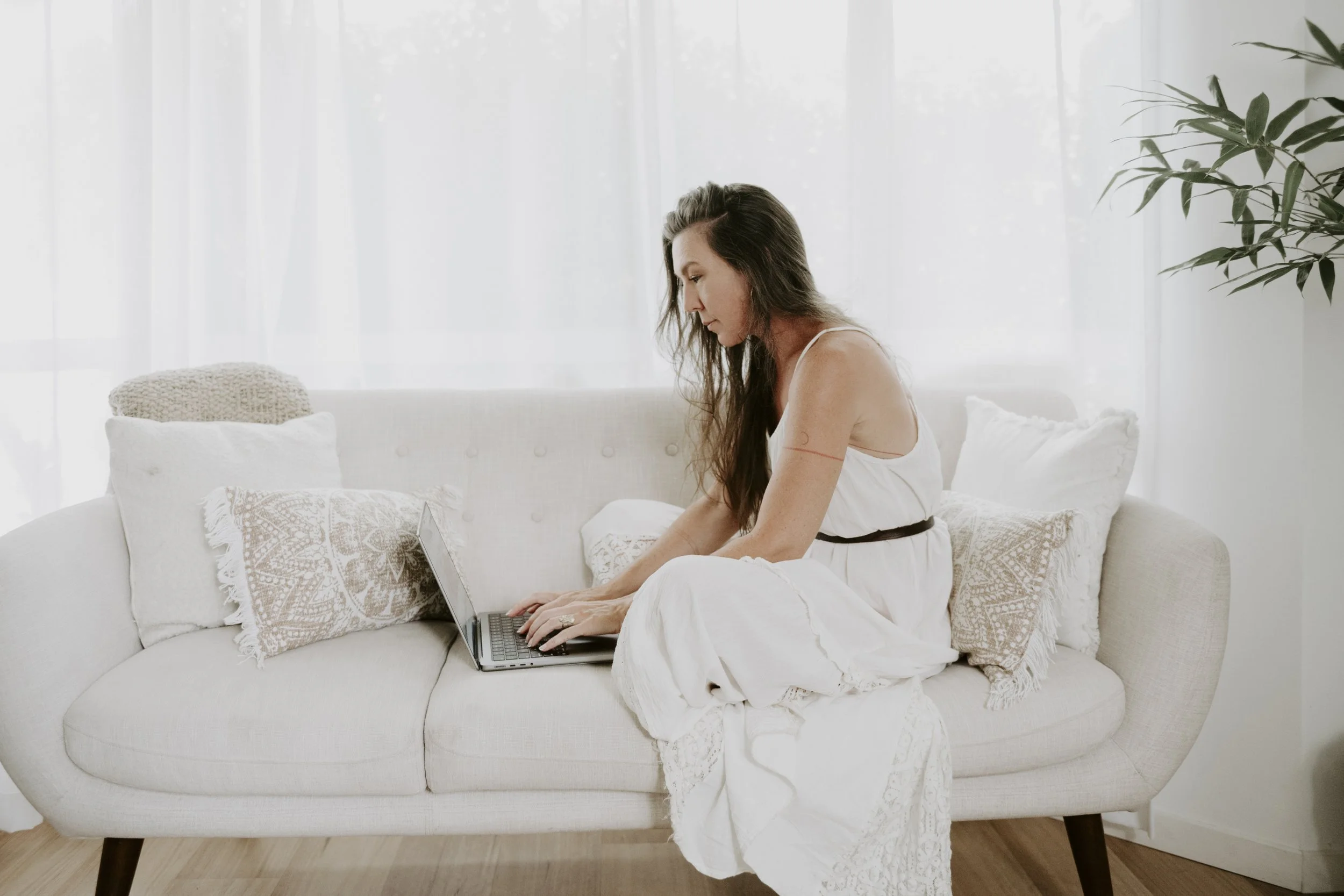 Mariko with long dark hair sitting on a white couch, working on a laptop, in a bright room with white curtains and a large leafy green plant nearby.