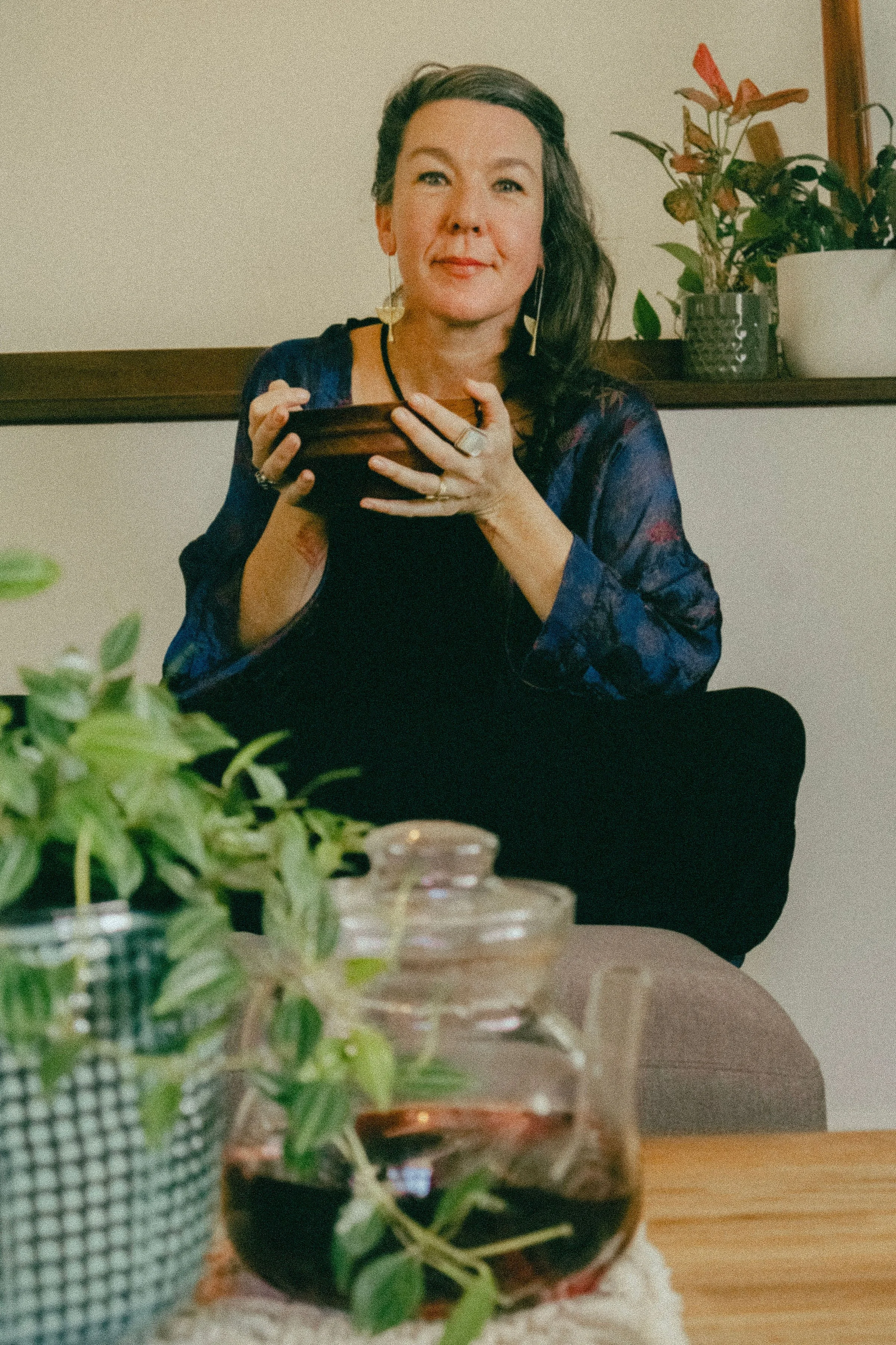 A woman with dark hair and earrings, wearing a blue and purple top, sitting at a table with a plant in a black pot in front of her, holding a wooden bowl, and smiling, with a plant and picture frame in the background.