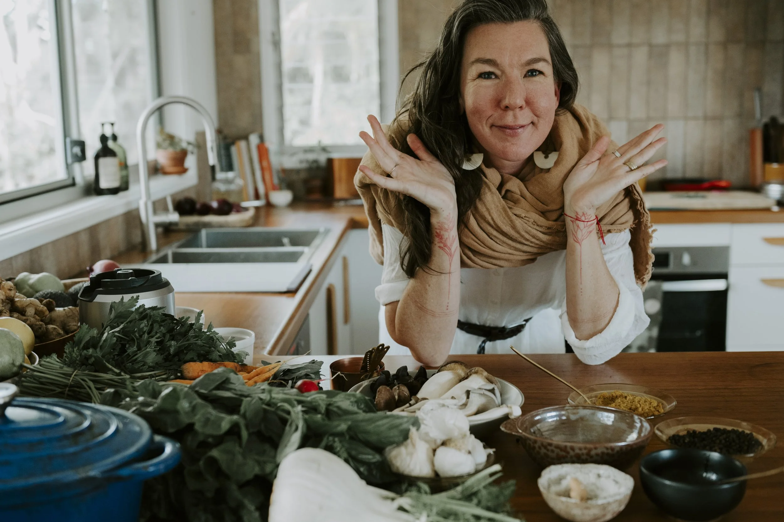 Mariko the midwife smiles at the camera while leaning on a kitchen counter filled with various vegetables, herbs, and bowls of food. The kitchen has large windows and a rustic decor.