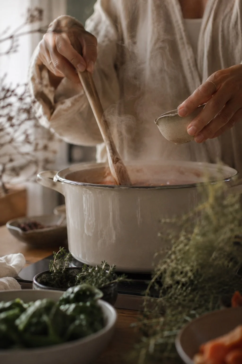 Mariko the midwife stirring a pot of steaming food in a kitchen, with herbs and bowls on the table.