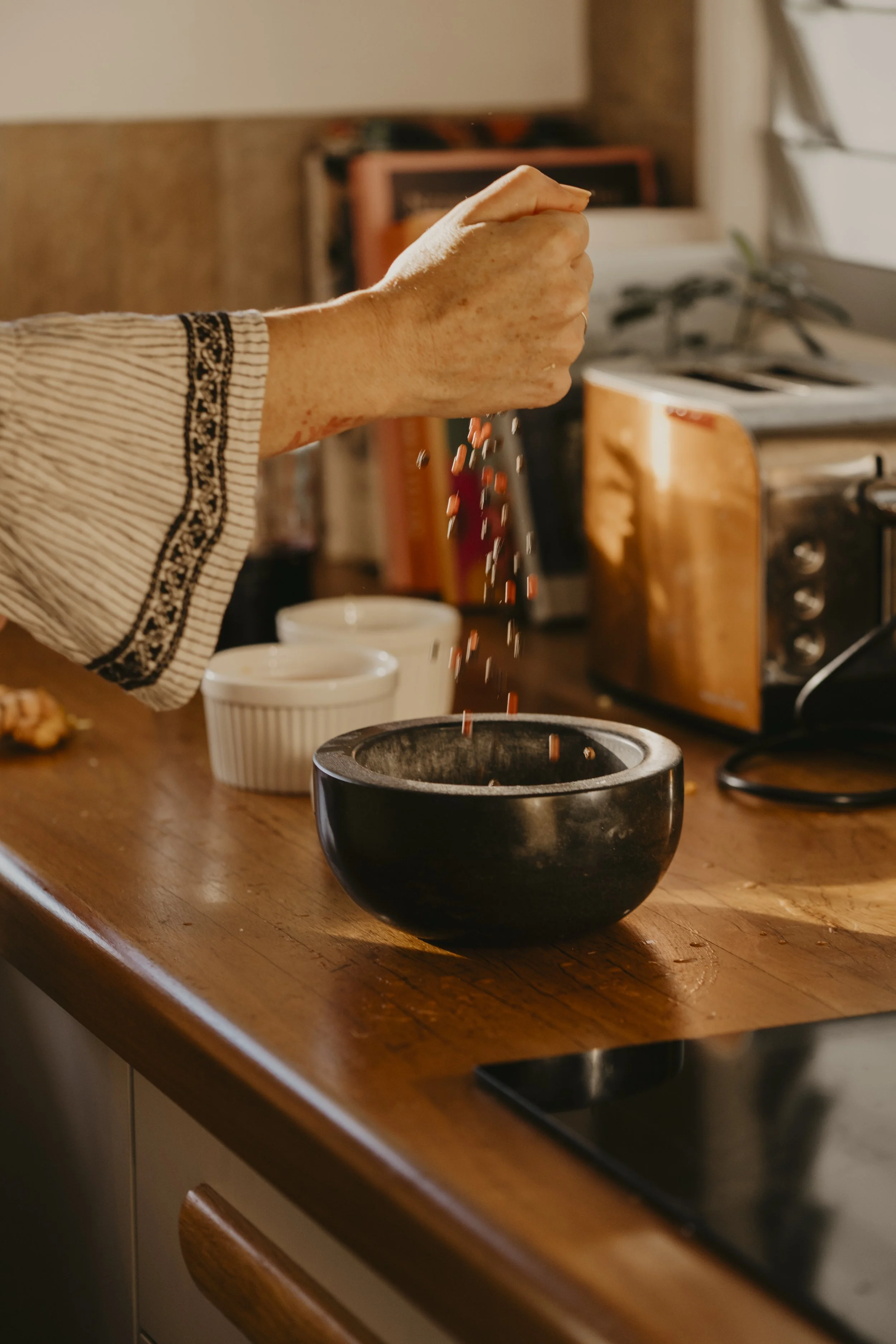 Marikos hand is sprinkling small red spices into a black bowl on a wooden kitchen counter. In the background, there are white bowls, a toaster, and a stove.