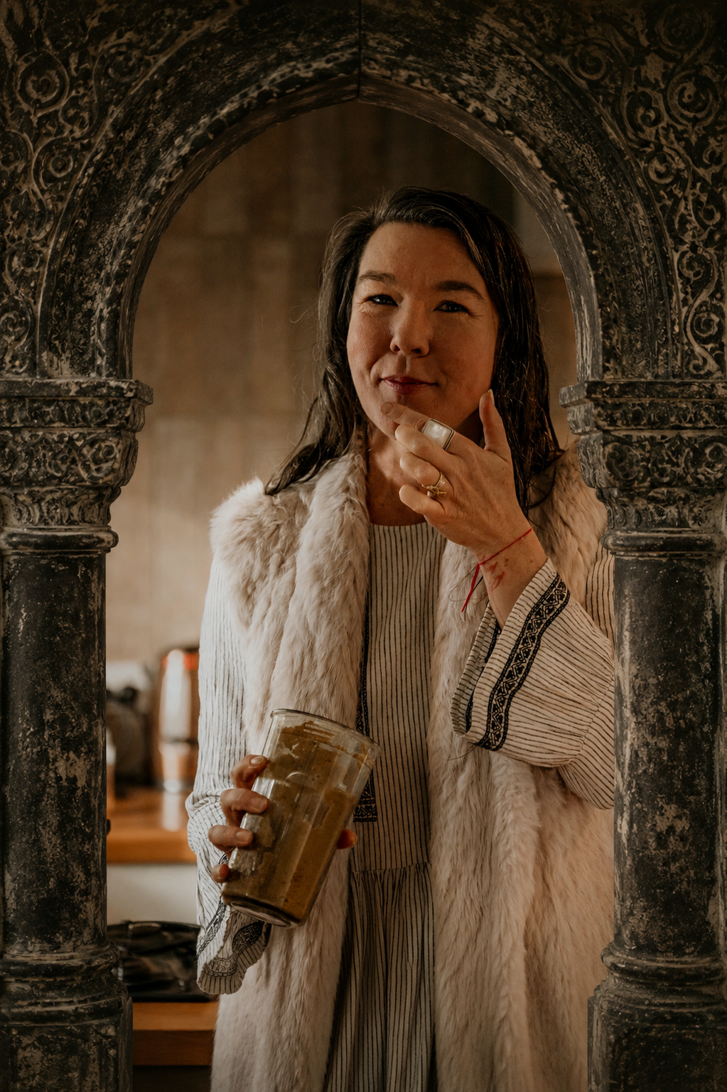 A woman with wet hair standing in front of an ornate stone archway, holding a glass of iced coffee and touching her chin with her other hand, dressed in a striped dress and a faux fur vest.