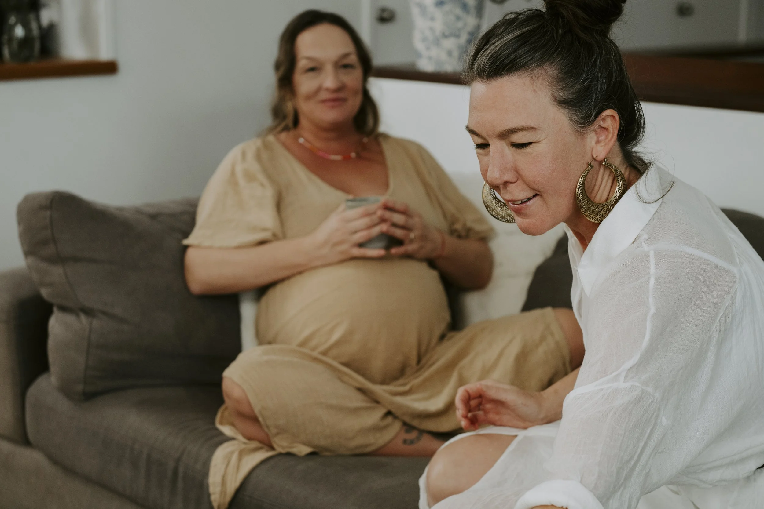 Two women sitting on a gray couch. One woman is pregnant, wearing a beige dress, holding a mug, and smiling. The other woman, wearing a white shirt with rolled sleeves and large hoop earrings, is leaning forward and smiling.