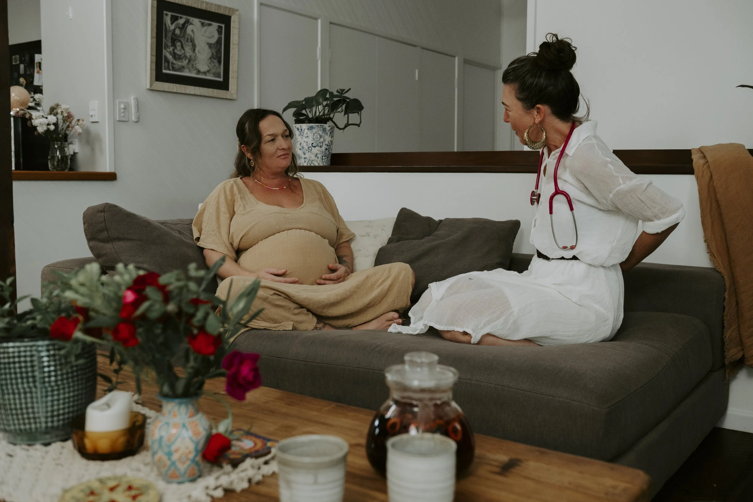 A pregnant woman sitting on a couch, looking concerned, while a healthcare professional, possibly a nurse or doctor, standing and talking to her. The scene appears to be in a living room with a table in the foreground decorated with vases, flowers, and drinks.