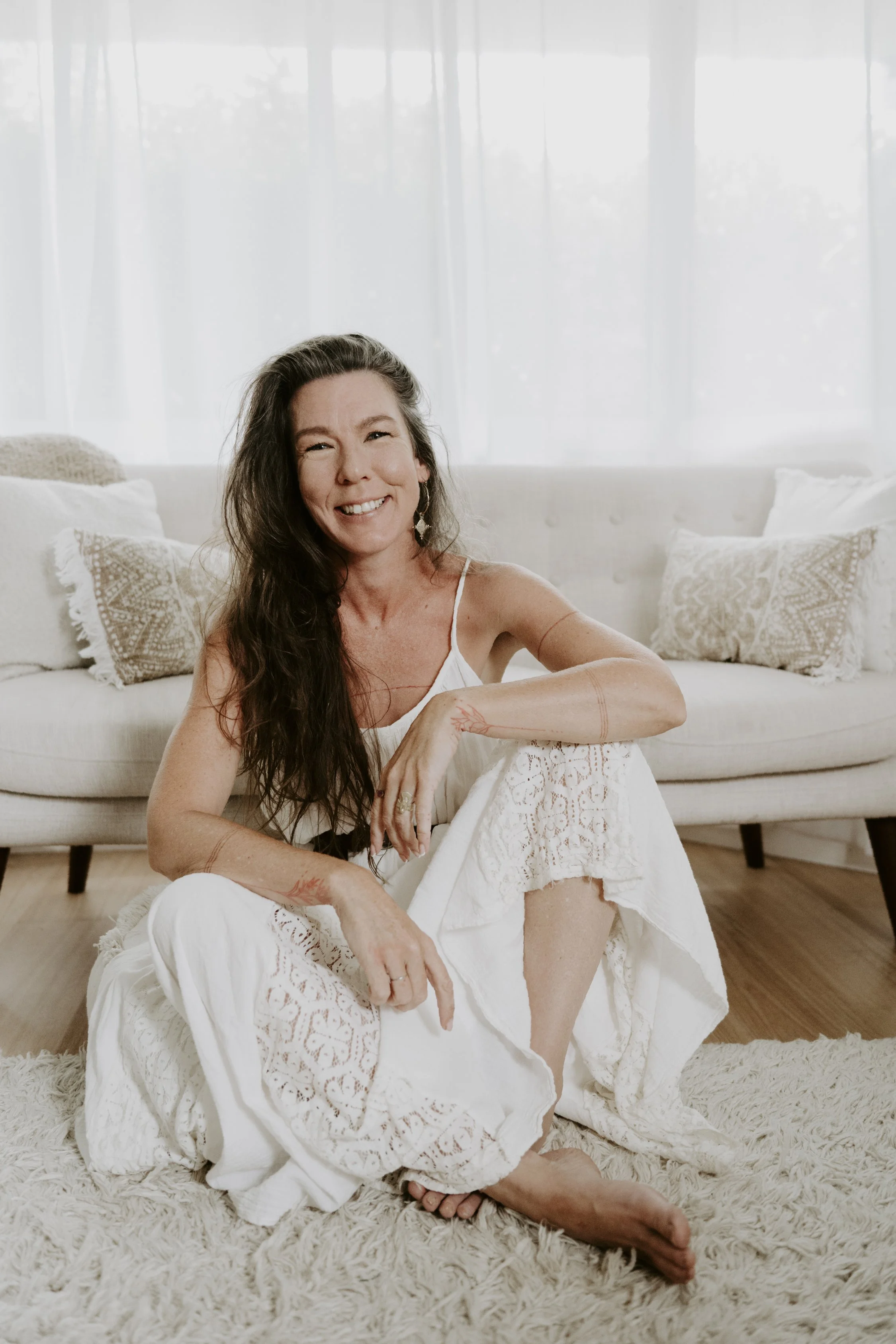 Woman sitting on a fluffy carpet in a bright room with sheer curtains, smiling at the camera, wearing a white dress with lace details.