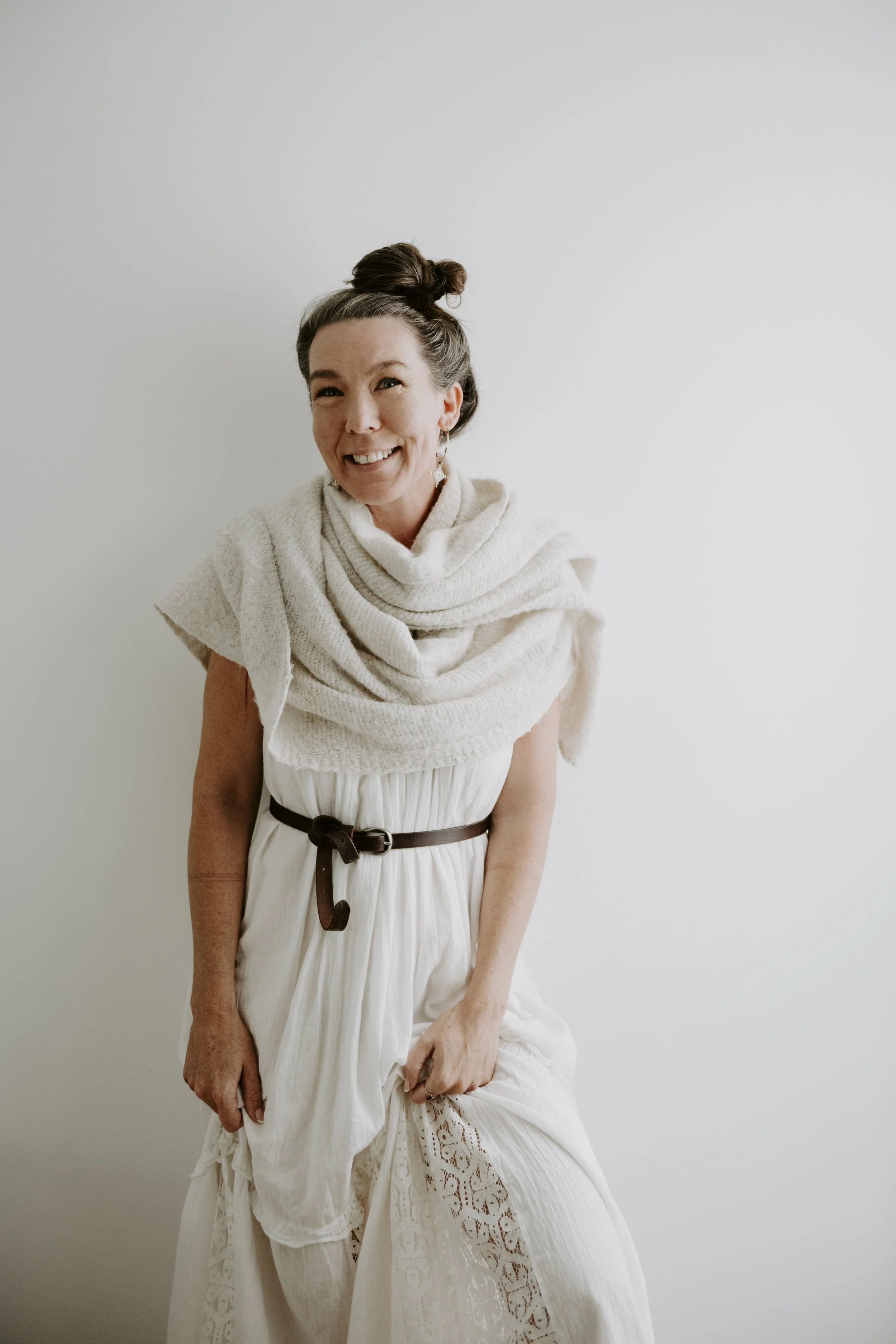 Mariko with gray hair in a topknot, smiling, dressed in a white scarf or shawl, standing against a plain light gray wall.