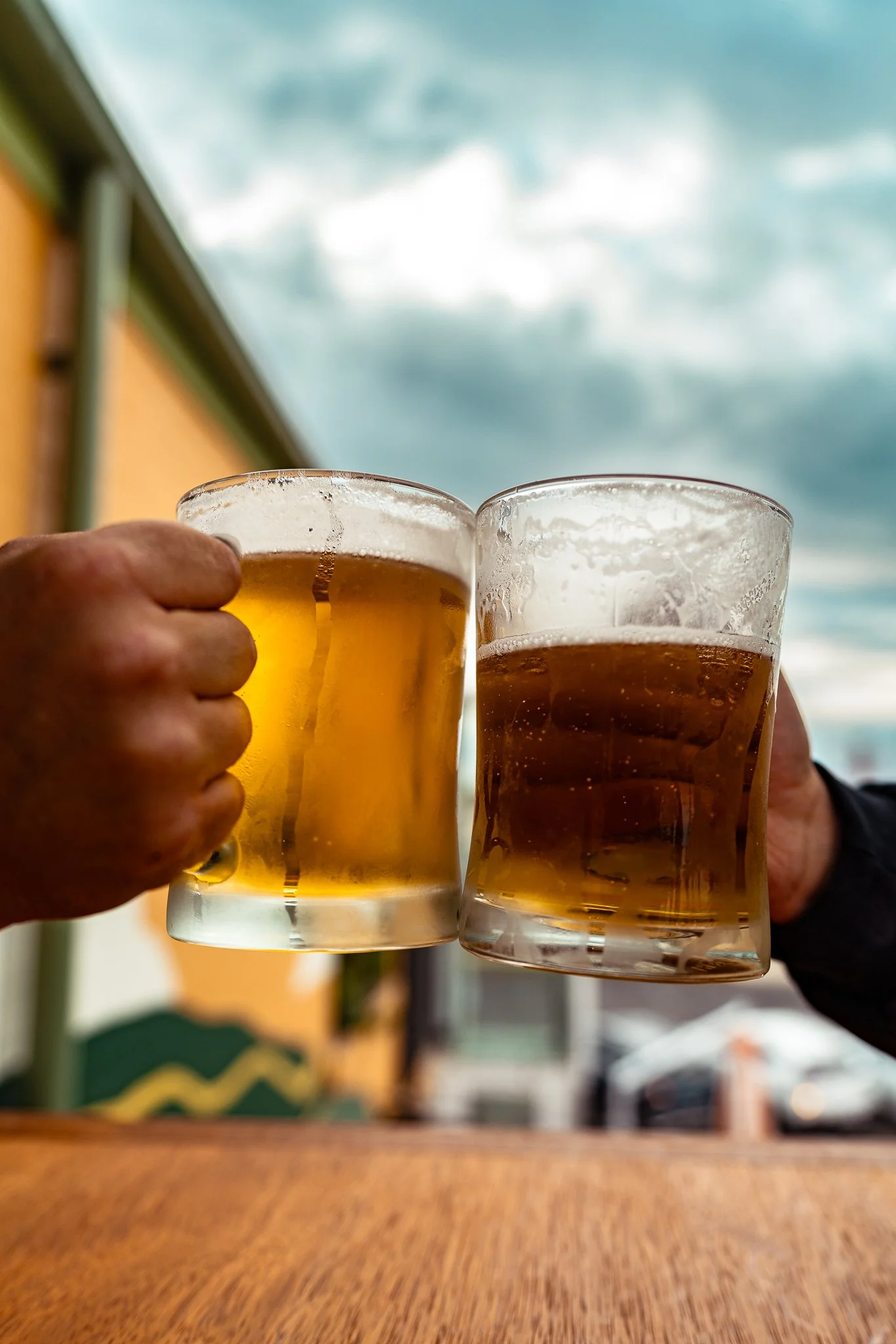 Two people clinking beer glasses outdoors against a cloudy sky.