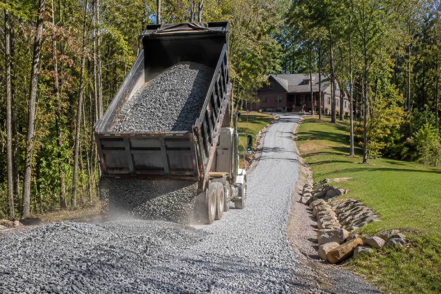 A dump truck unloading gravel on a residential driveway lined with rocks, with trees and a house in the background.