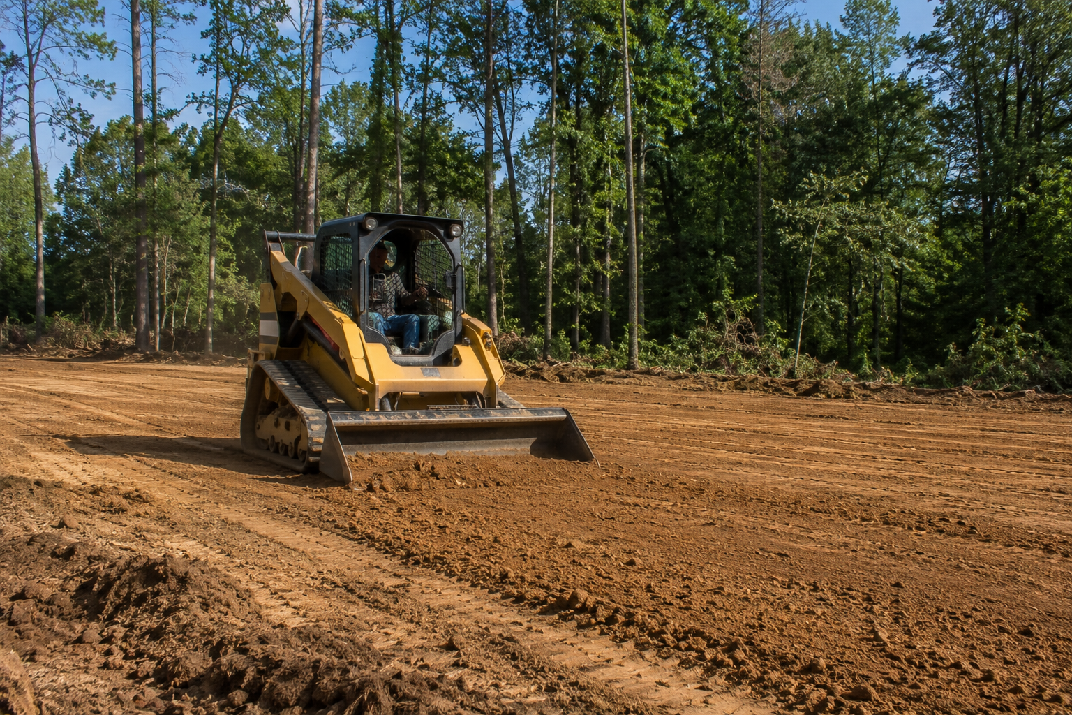 A person operating a yellow bulldozer on a cleared dirt area surrounded by trees.
