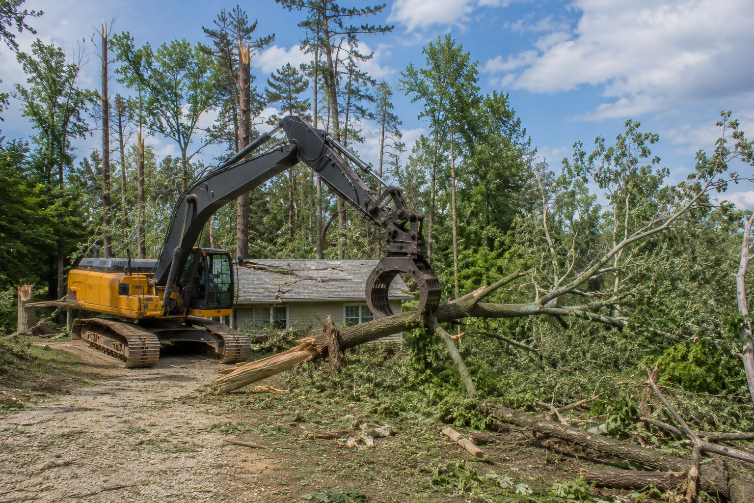 A yellow excavator tearing down a fallen tree in front of a house with a damaged roof, surrounded by green trees and a partly cloudy sky.