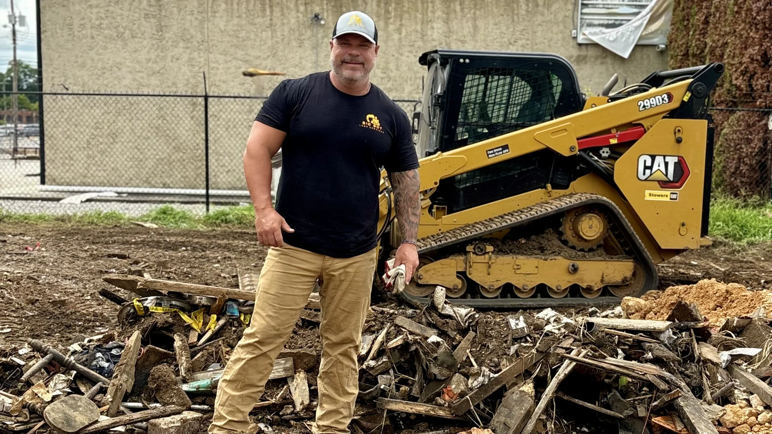 A man standing in front of a small yellow excavator on a construction site, surrounded by dirt and debris. The man is wearing a black t-shirt, khaki pants, and a gray cap, and holding a pair of work gloves.