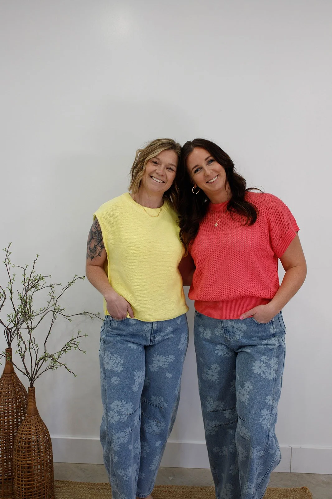 Two women standing close together, smiling, in front of a plain white wall. One woman is wearing a yellow sleeveless top and the other is wearing a red short-sleeve top. They are both wearing blue jeans with floral patterns and have their hands in their pockets. There is a small potted plant with thin branches on the left side of the image.