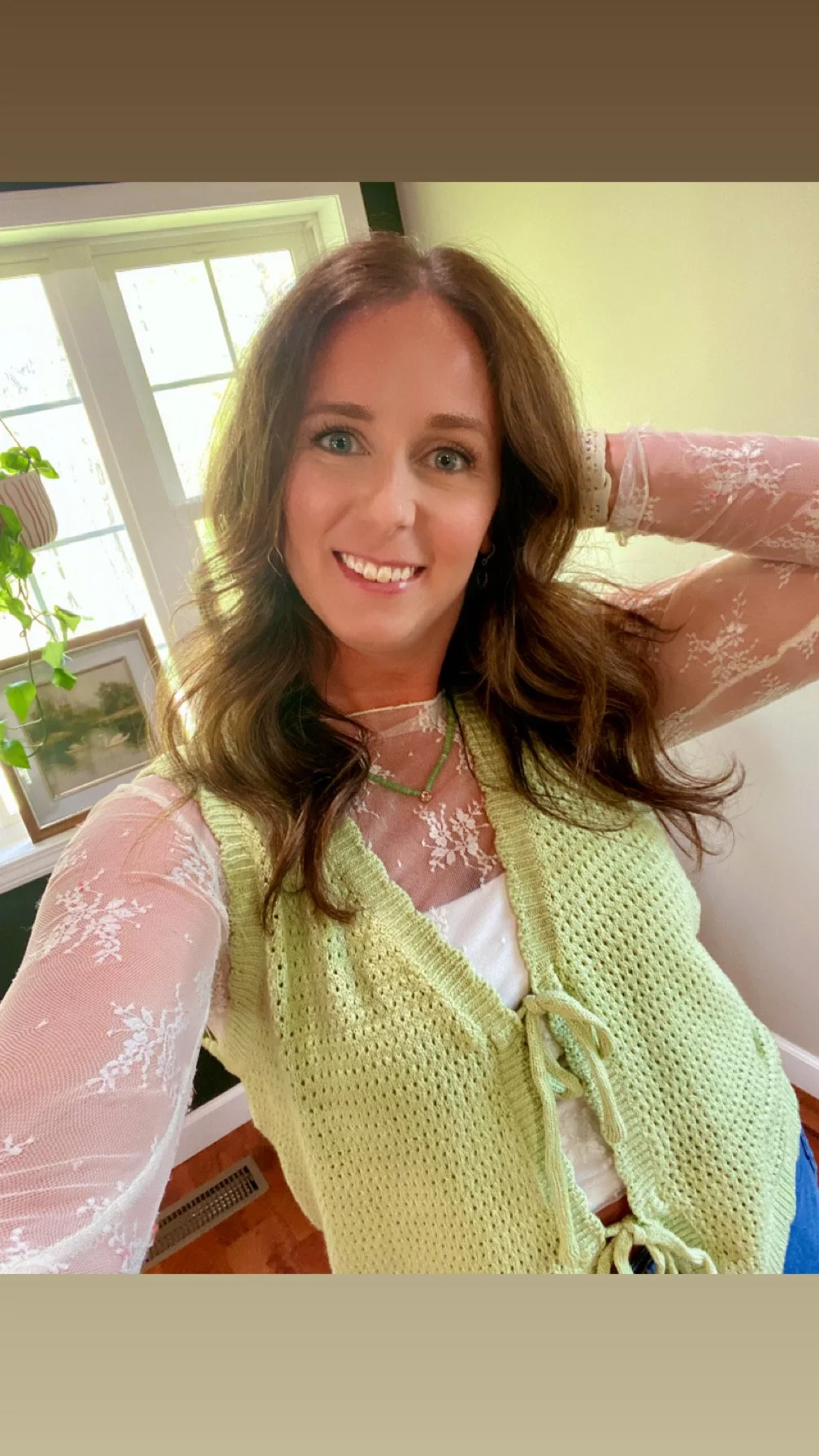 A woman with wavy brown hair smiling indoors, wearing a pink lace top and a yellow crochet vest, standing near a window with green walls.