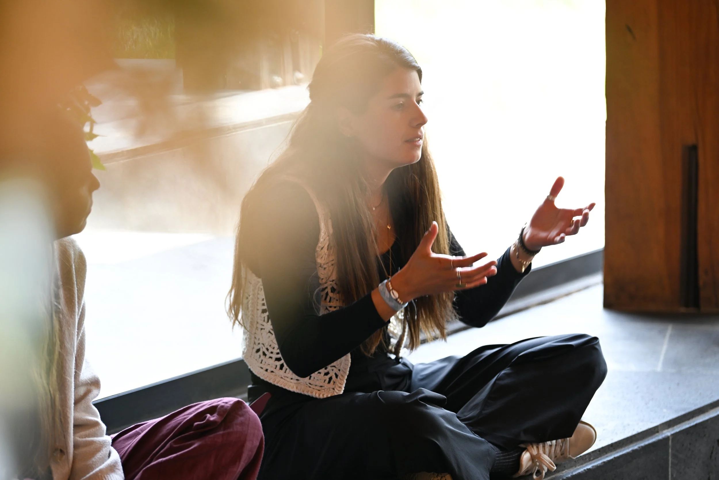 A woman with long hair sitting cross-legged on the floor, talking with expressive hand gestures, next to a young girl with glasses and curly hair, both sitting near a wooden wall with sunlight coming through a window.