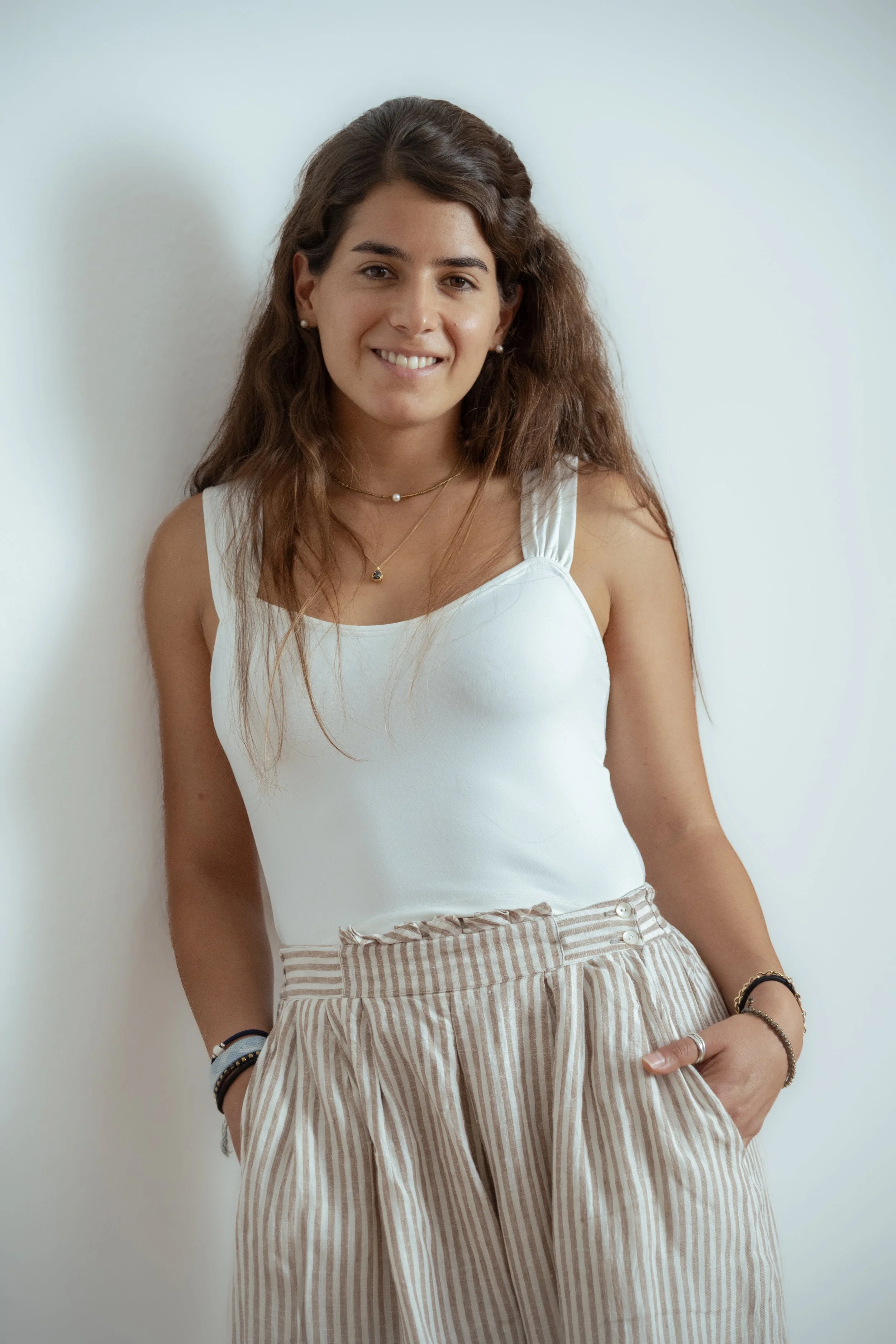 A young woman with long wavy brown hair, wearing a white sleeveless top, striped beige and white pants, and accessorized with layered necklaces, earrings, and bracelets, standing against a plain white wall.