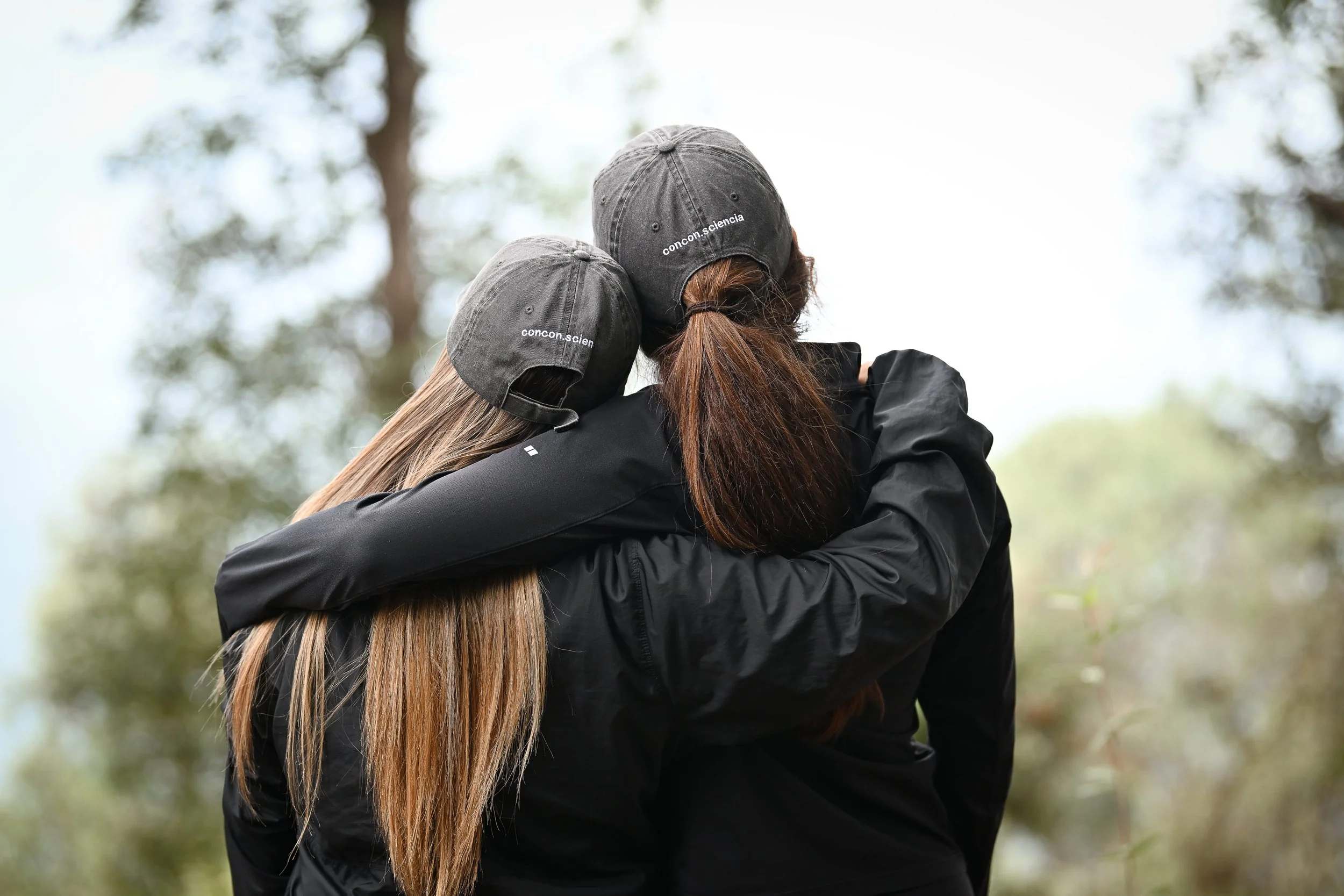 Two people embracing outdoors, one with long hair and the other with shorter hair, both wearing black jackets and gray baseball caps with the text 'concon.ciencia' on them, surrounded by trees and natural greenery.