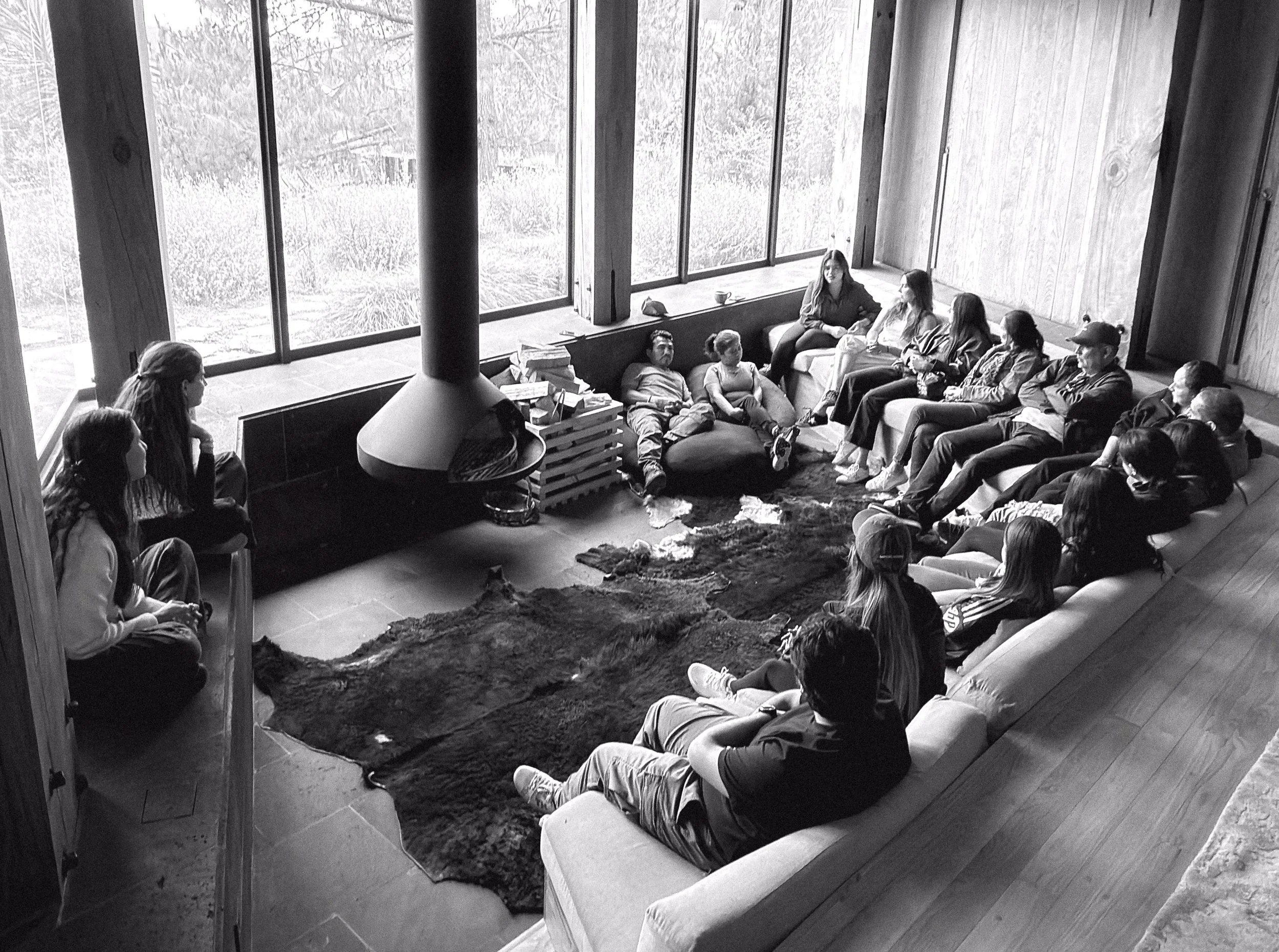 A group of people seated in a living room engaged in conversation, with large windows showing an outdoor view.