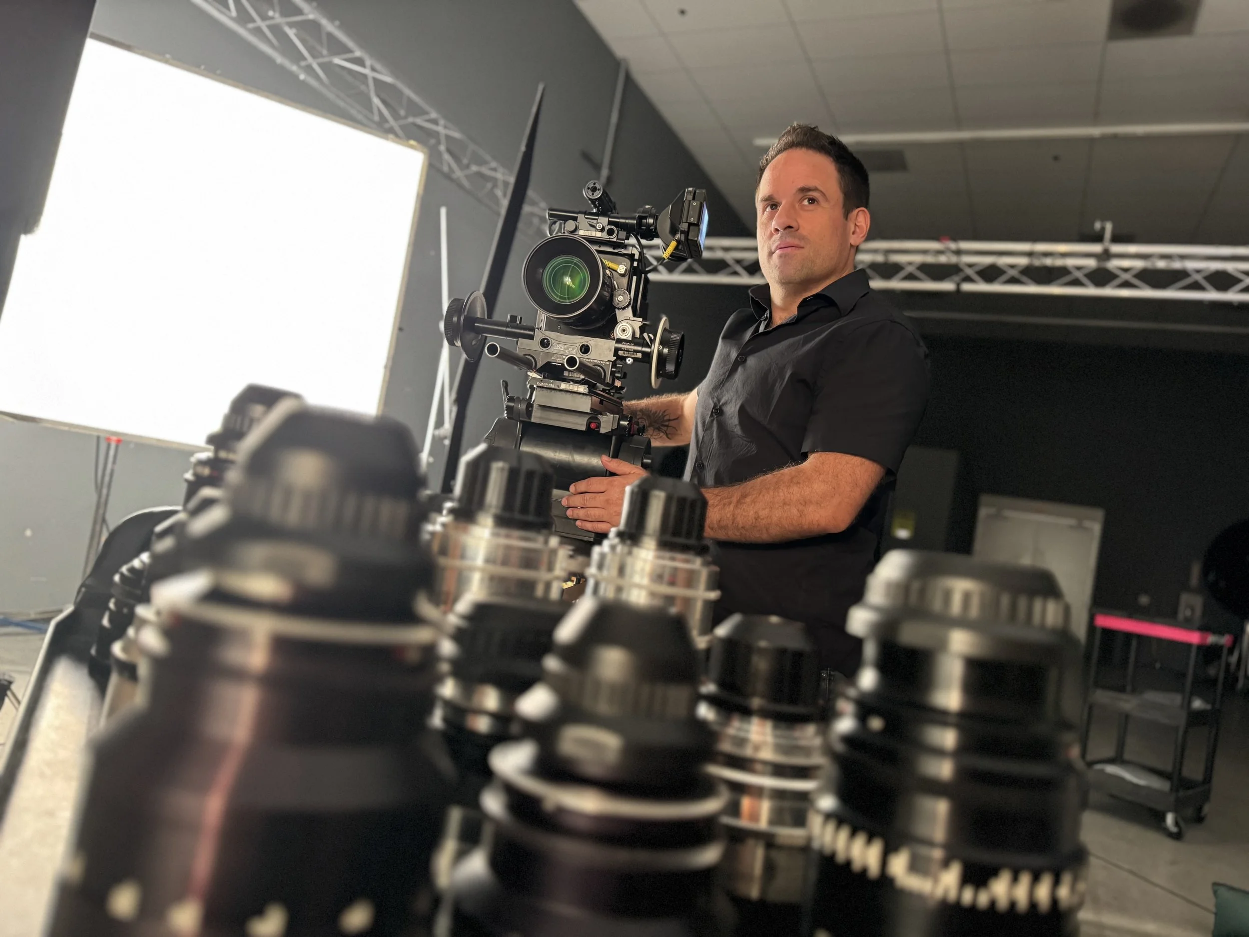 A man in black shirt operating a large camera mounted on a rig, with lighting equipment and stage trusses in the background, in a studio or technical production environment.