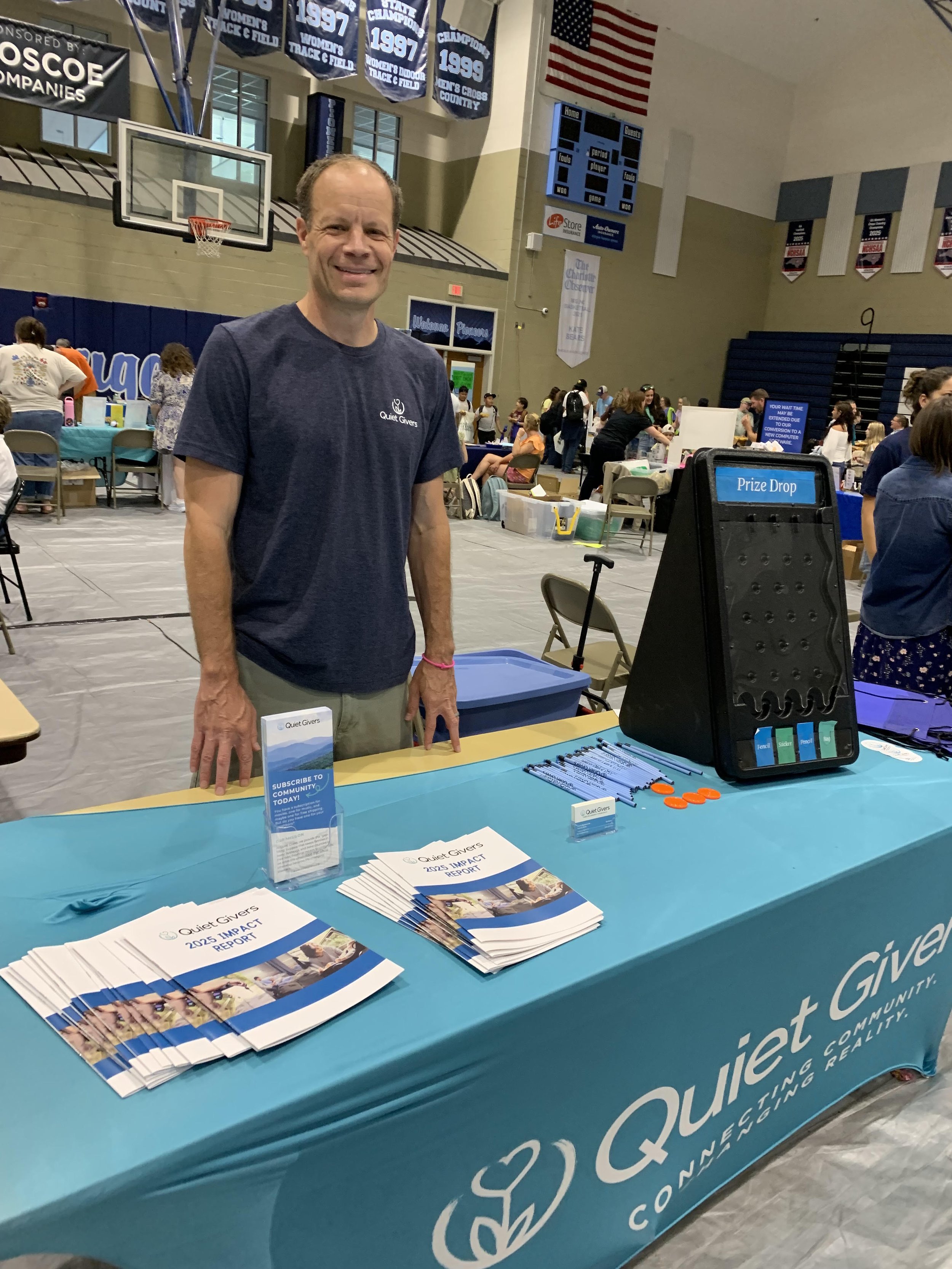 A man standing behind a table with blue tablecloths at an indoor event. The table displays flyers and brochures for Quiet Givers, a community organization. The man is smiling and wearing a dark blue T-shirt with the Quiet Givers logo. There are people and booths in the background.