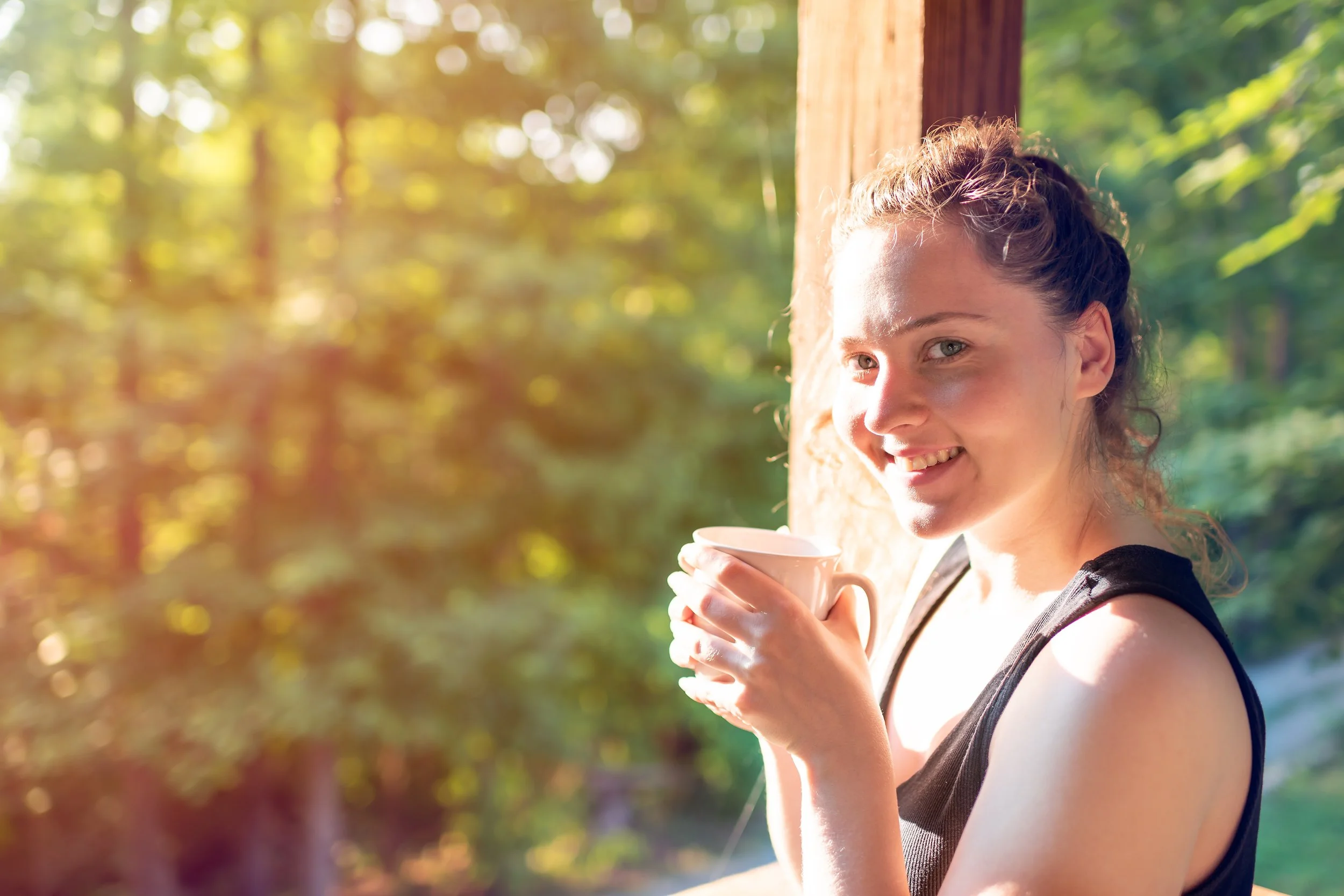 A woman with curly hair holding a white mug and smiling outdoors in a wooded area with sunlight filtering through the trees.