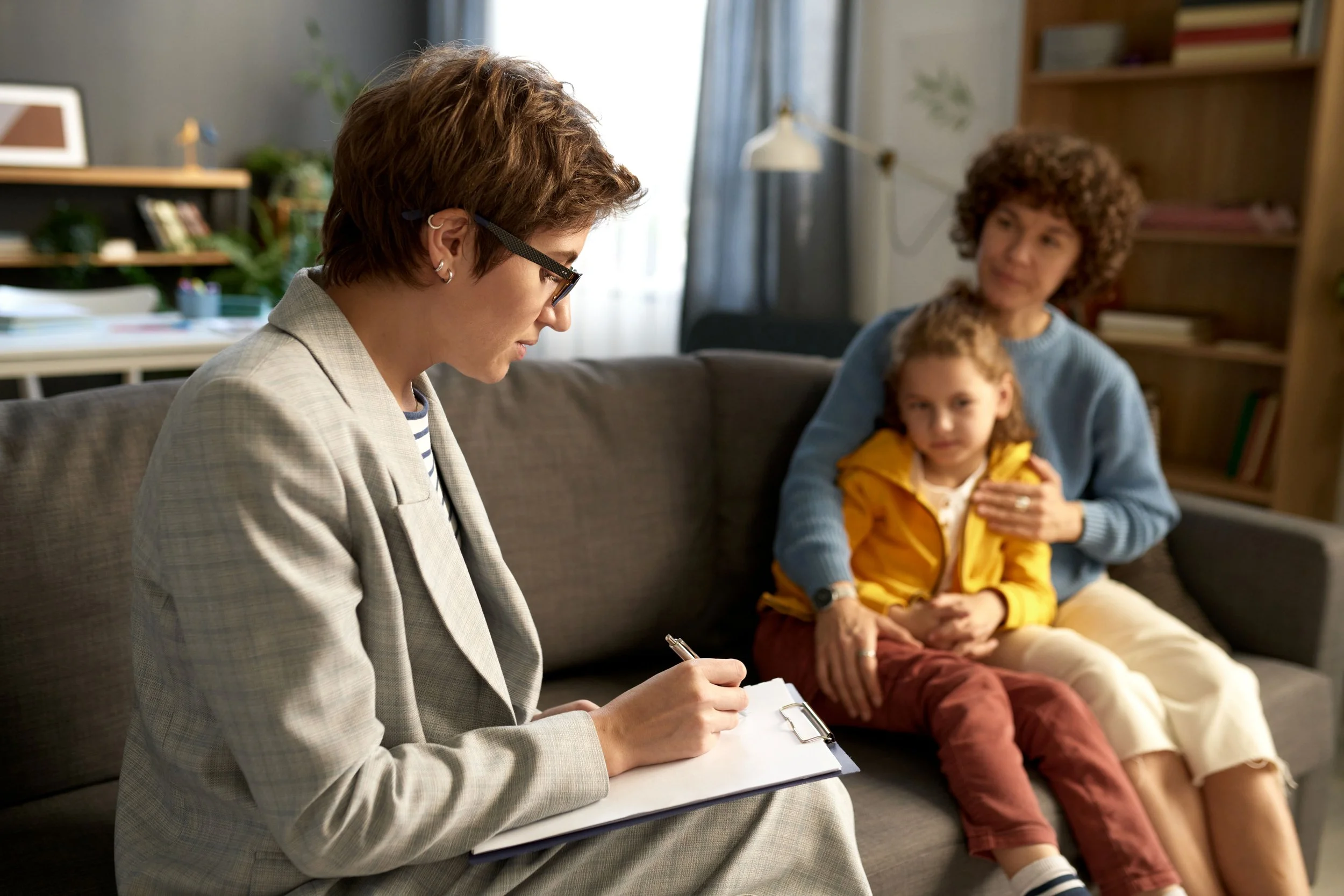 A woman with short brown hair, glasses, and earrings wearing a plaid blazer writing on a clipboard, with a woman and a child sitting on a couch in the background