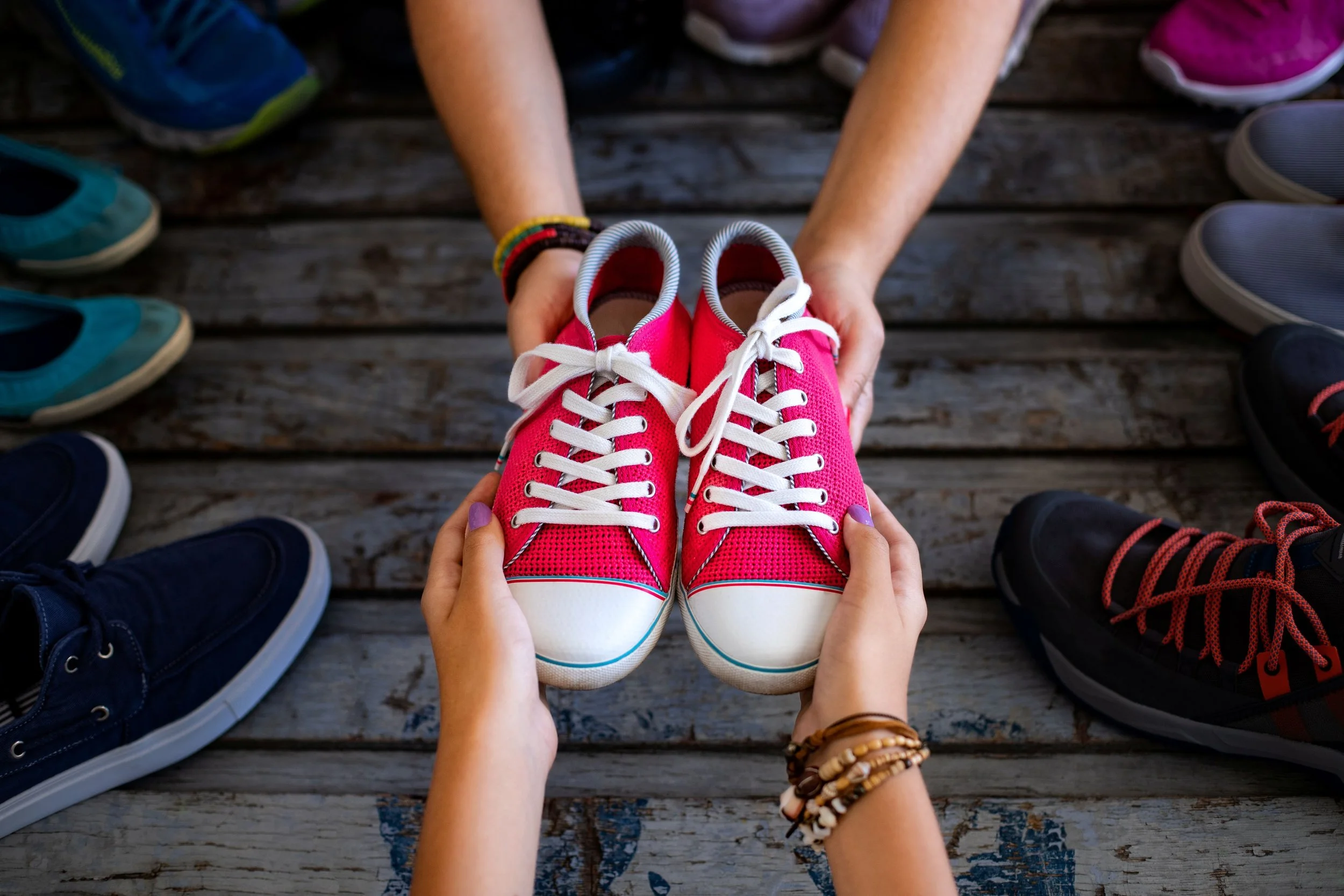 Person holding pink sneakers with white laces, surrounded by various pairs of shoes on a wooden surface.