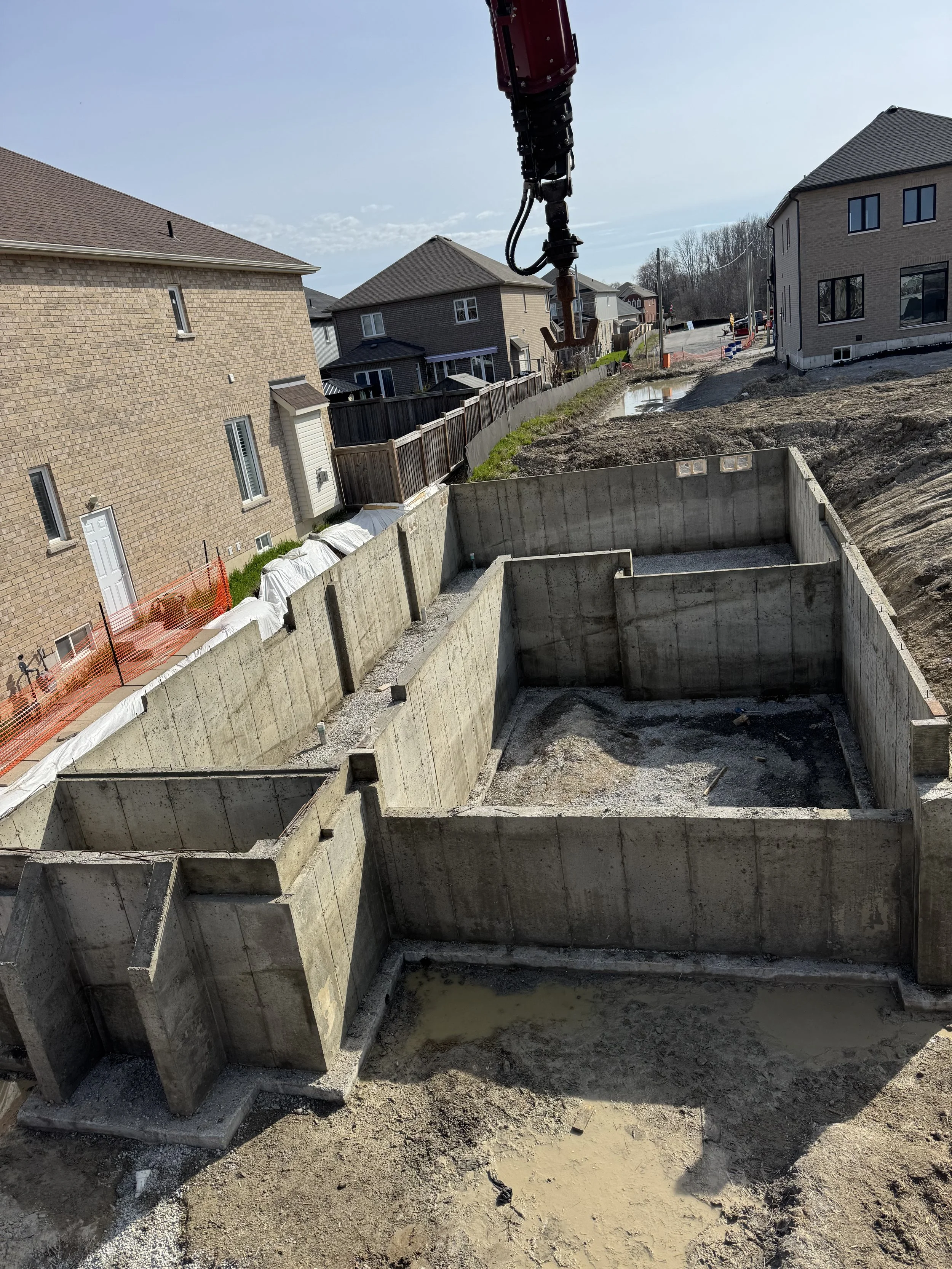 Concrete foundation forms for a building at a construction site, with surrounding dirt, nearby houses, and a construction crane's arm visible.