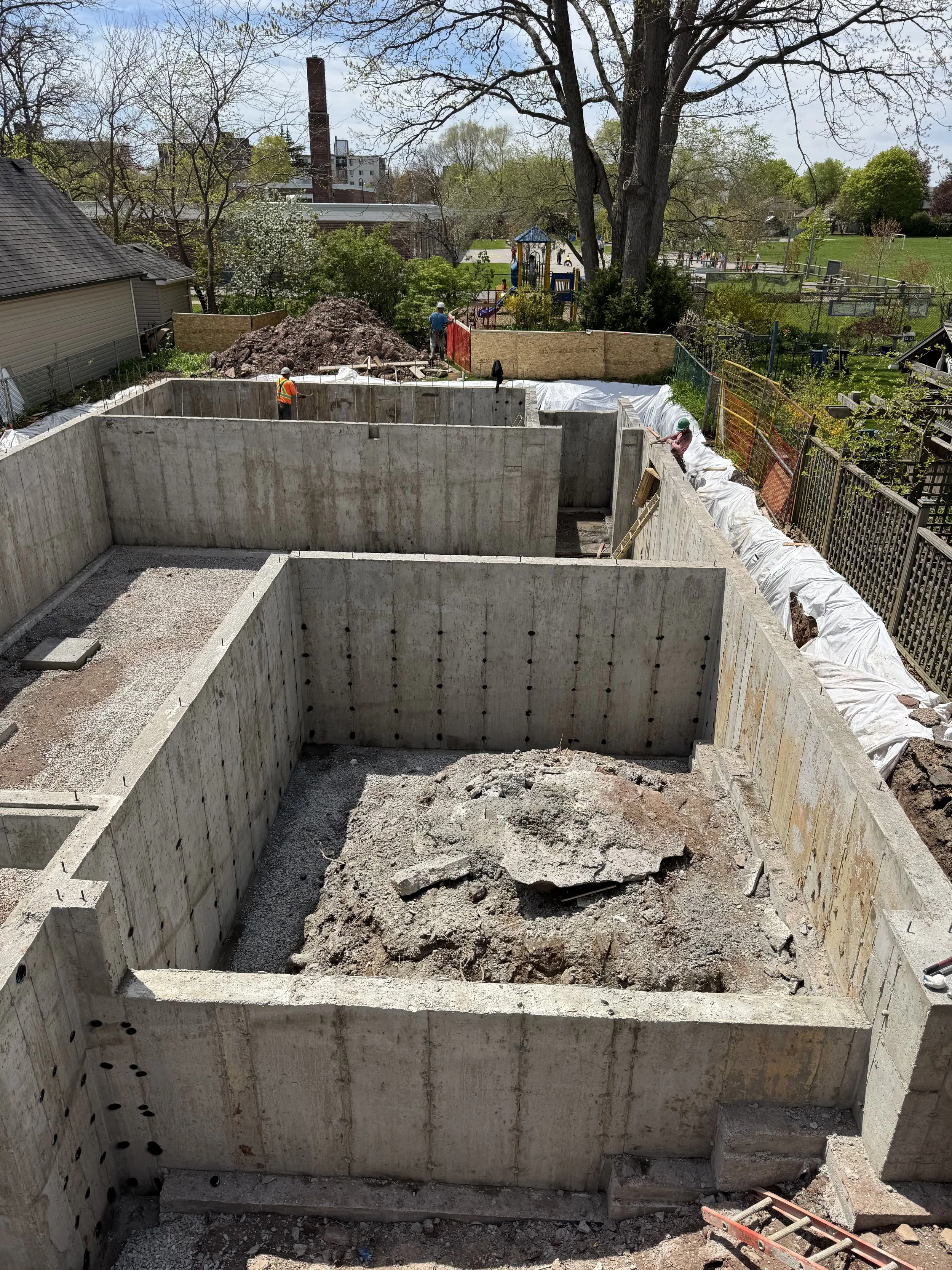 Construction site with concrete foundation walls and workers installing drainage or utility pipes, with a residential neighborhood and park in the background.