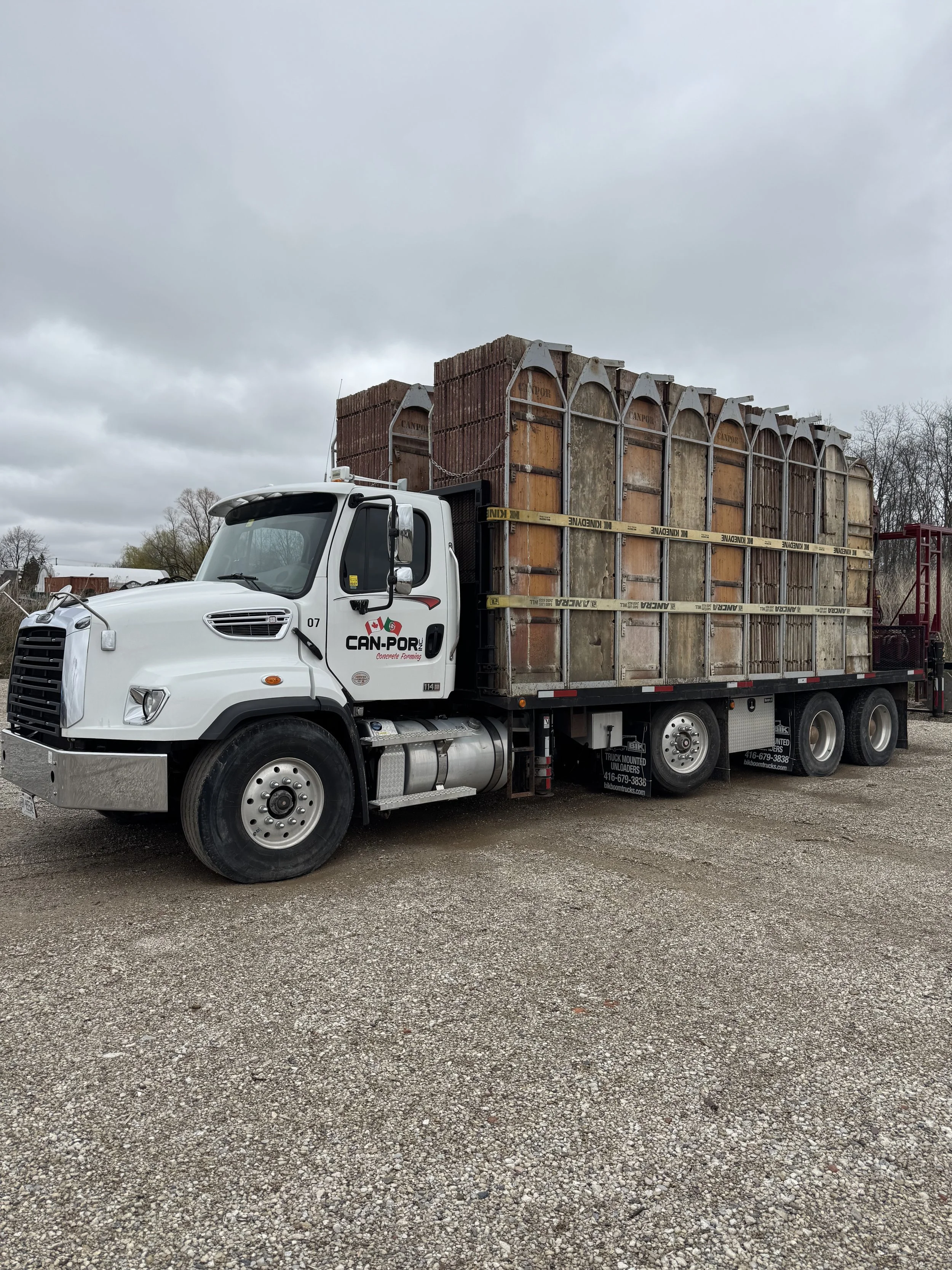 A large white truck carrying stacks of bricks loaded on a flatbed trailer, parked on a gravel lot on a cloudy day.