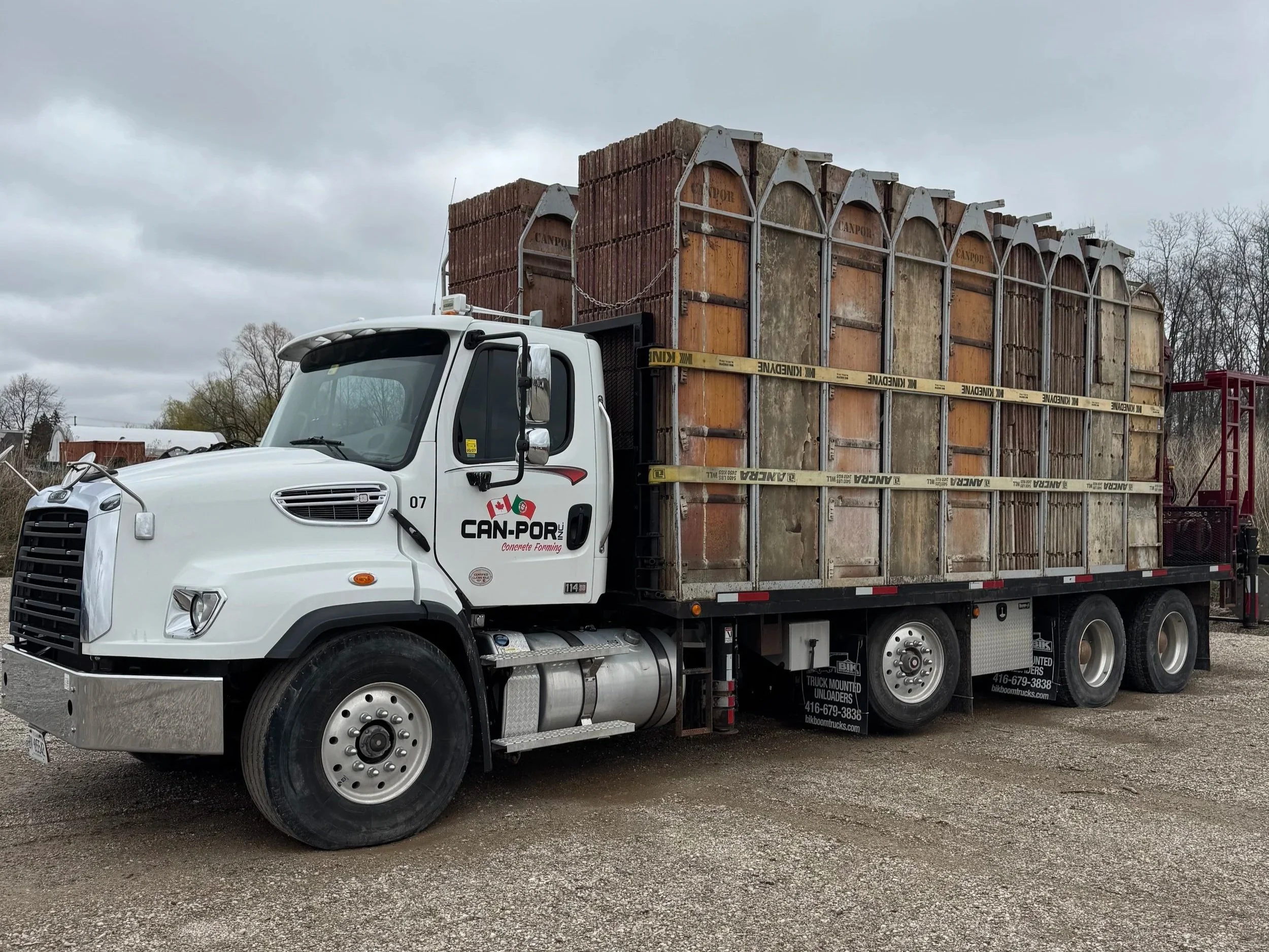 A white flatbed truck with the logo 'CAN-POR' on the door, carrying a large load of old, rusty wooden forms or panels, secured with yellow straps labeled 'KINDERV'. The background shows a cloudy sky and leafless trees.