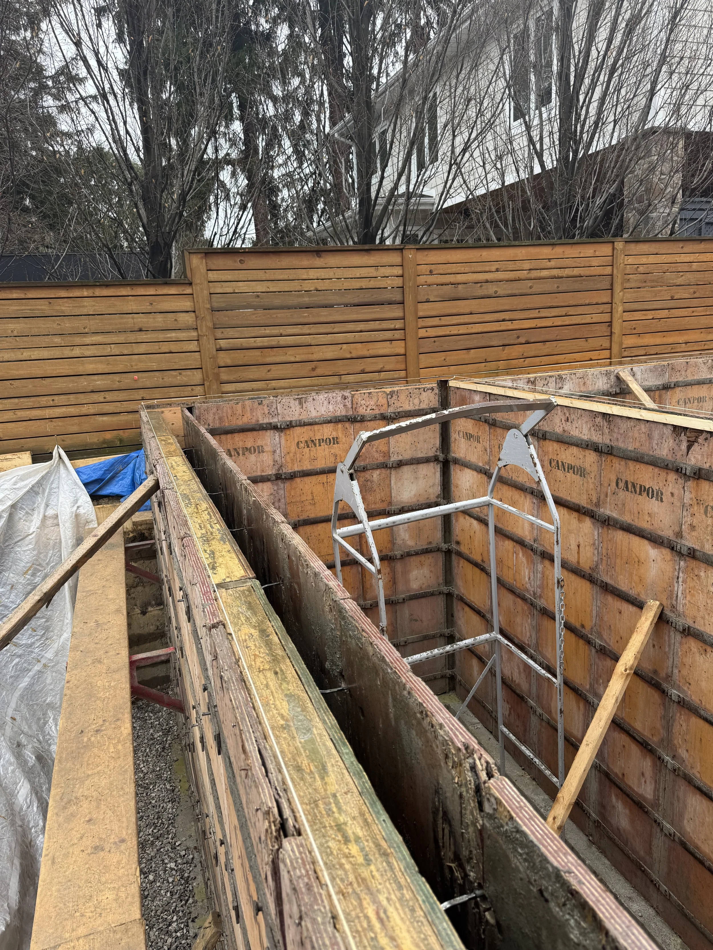 Construction site with exposed foundation walls and wooden framing. A wooden fence and some trees are visible in the background.