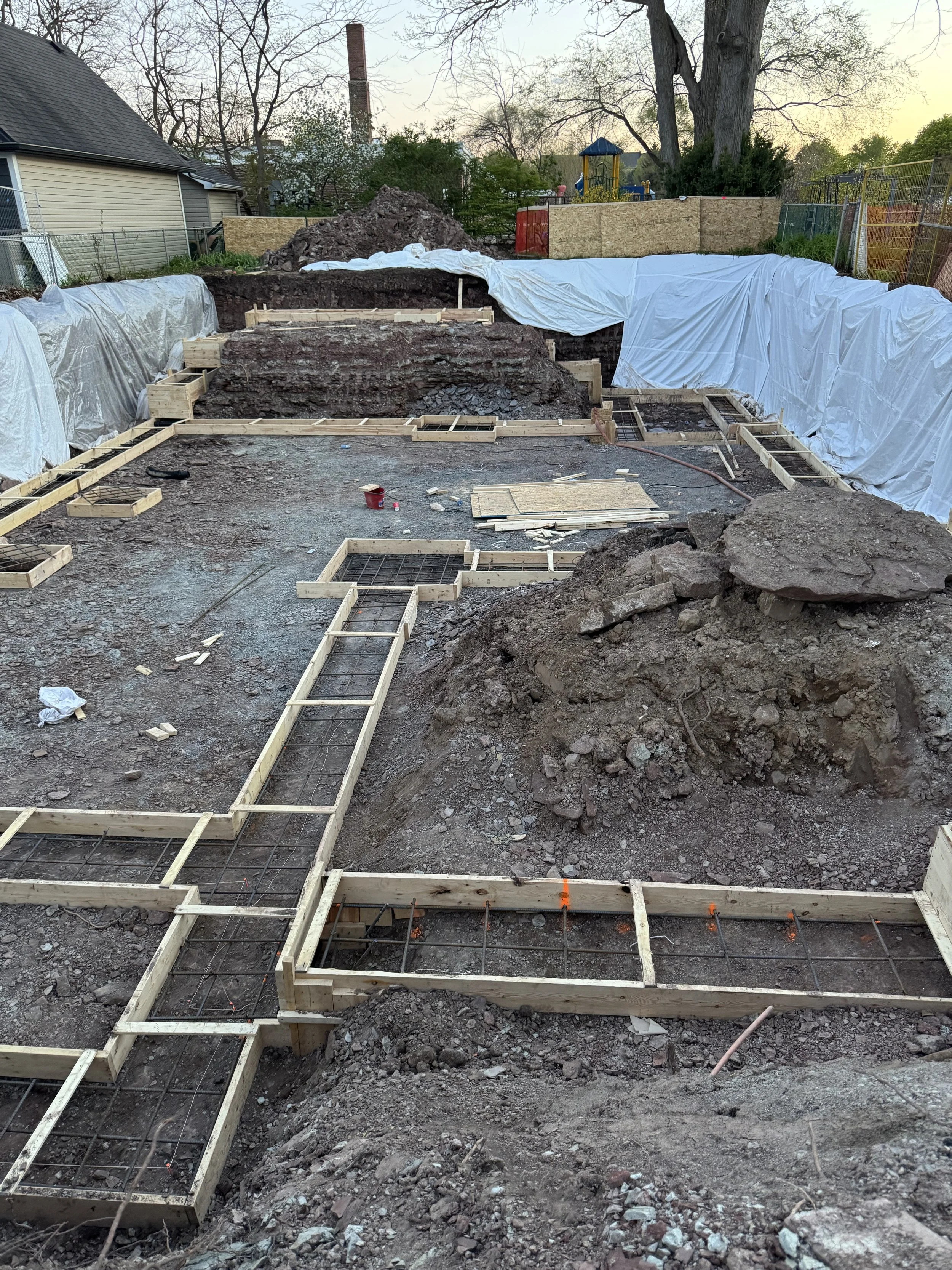 Construction site with wooden framework and rebar for a concrete foundation, dirt and rocks, and partially covered with white tarps, in an outdoor backyard.