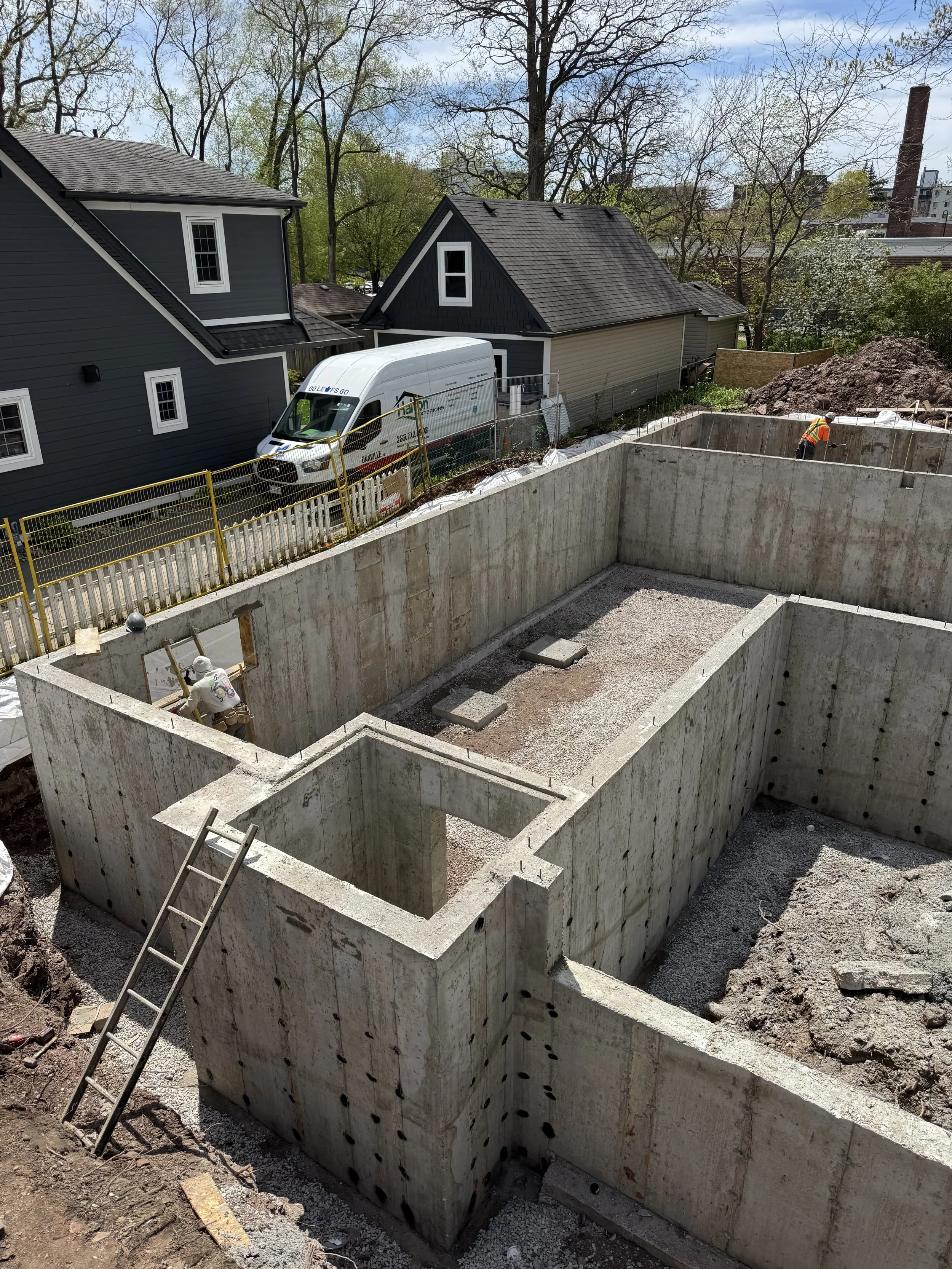 Construction site with excavated foundation for a building, with two workers and ladder, and residential houses in the background.
