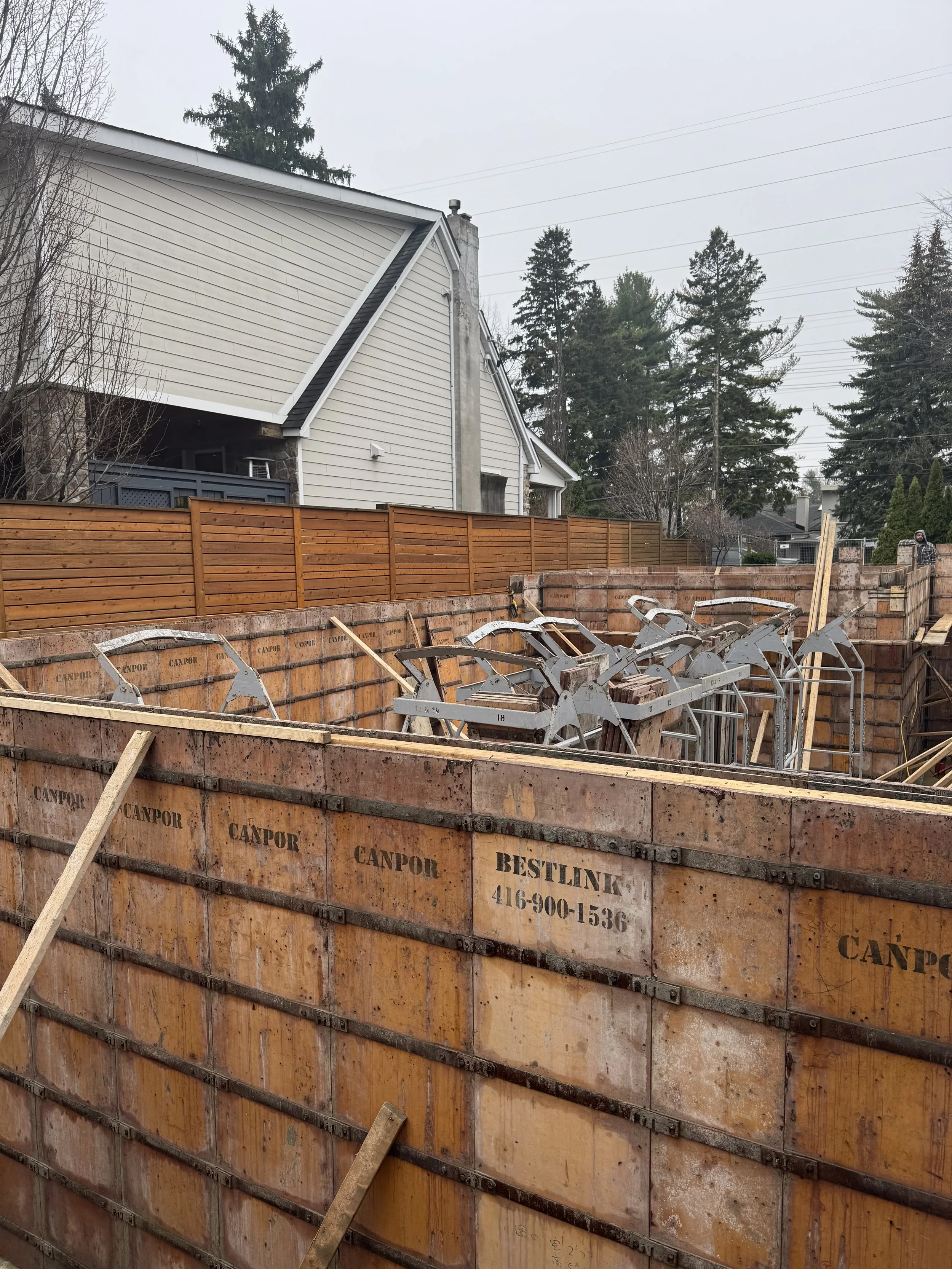 Construction site with a wooden foundation wall, metal framing supports, and a neighboring house with a yellow fence, trees, and a cloudy sky in the background.