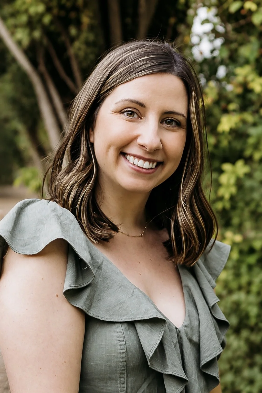 A young woman with shoulder-length brown hair and hazel eyes smiling outdoors, wearing a gray top with ruffled sleeves, in front of green trees and foliage.