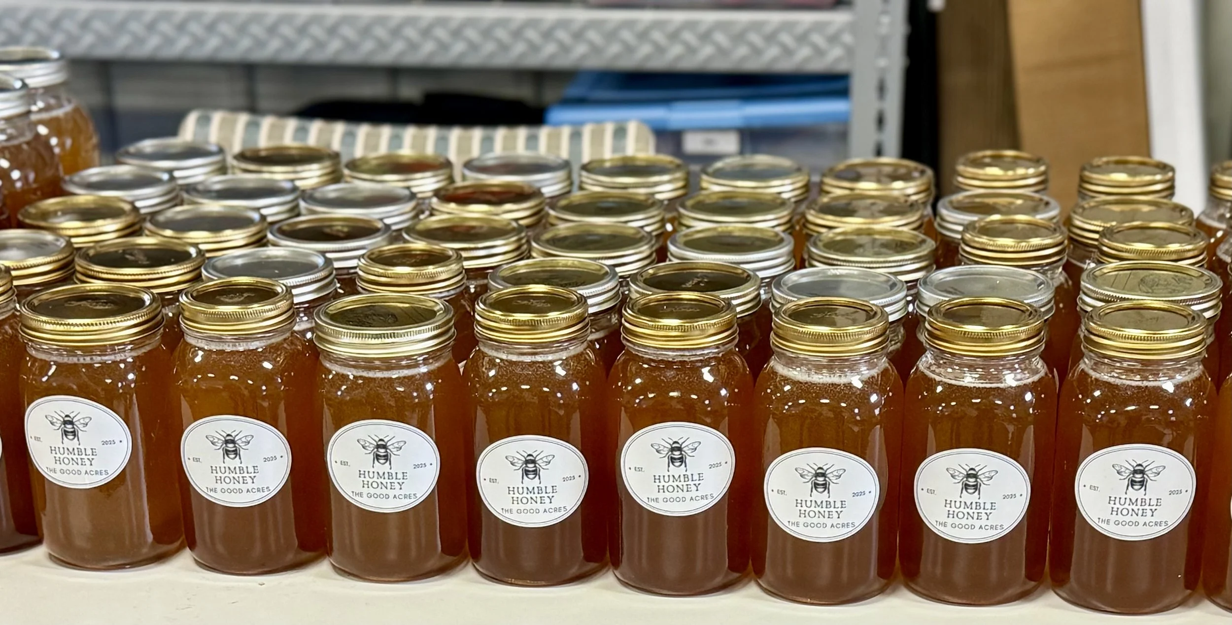 Row of honey jars with labels featuring a bee graphic and the text 'Humble Honey, The Good Acres' on a store shelf.