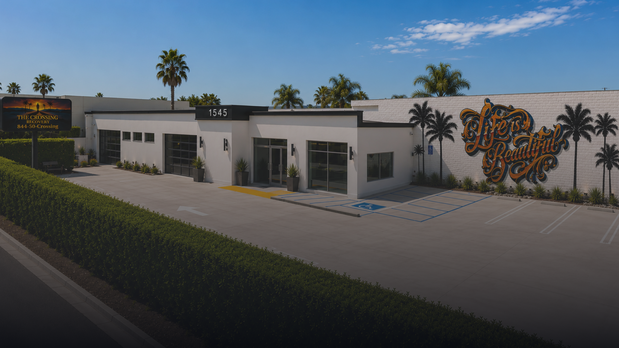Exterior of The Crossing Recovery, a drug and alcohol rehabilitation and outpatient addiction treatment center in Costa Mesa, California, featuring accessible parking, palm trees, modern facilities, and the “Life Is Beautiful” recovery mural.