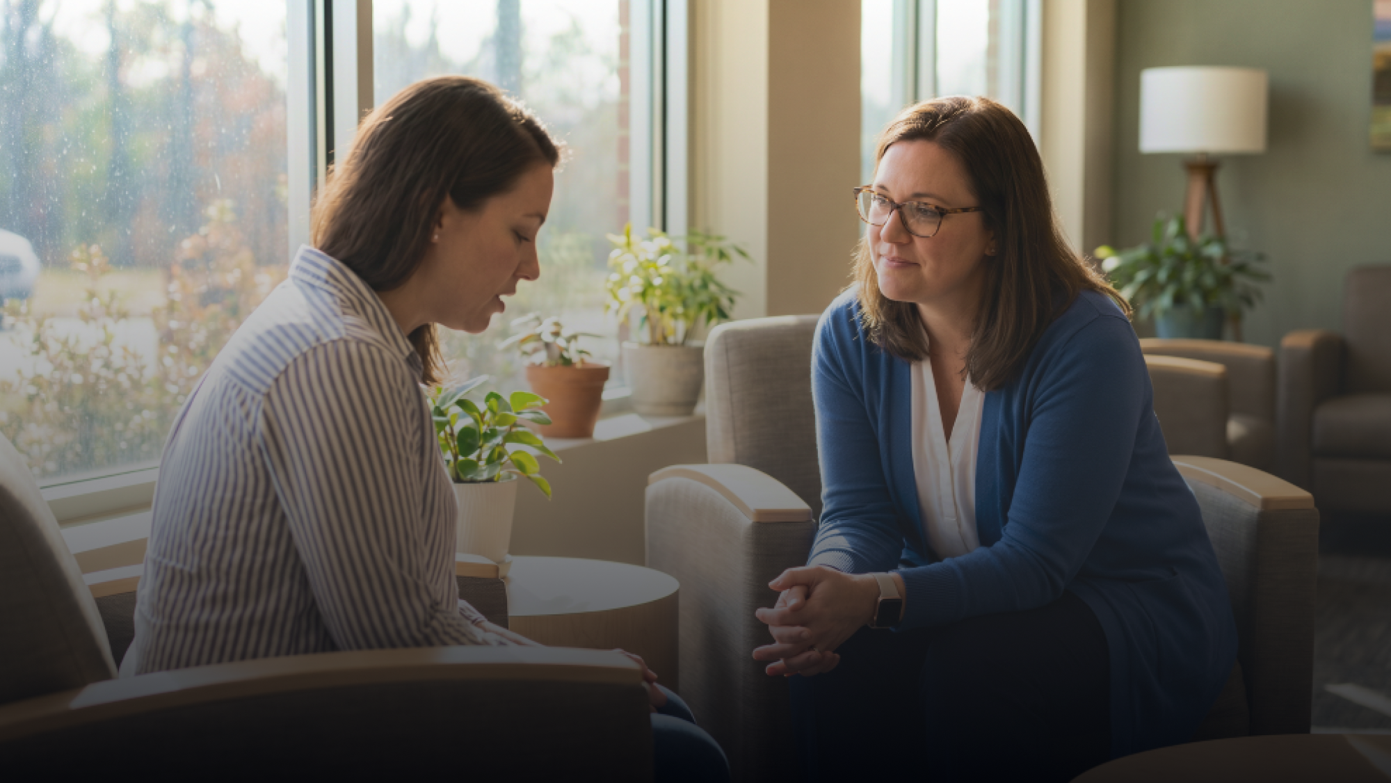 Two women sitting across from each other in a bright room with large windows, engaged in a discussion. One woman has shoulder-length dark hair and wears glasses and a blue blazer. The other woman has long, dark hair and wears a striped shirt. There are plants and a lamp in the background.