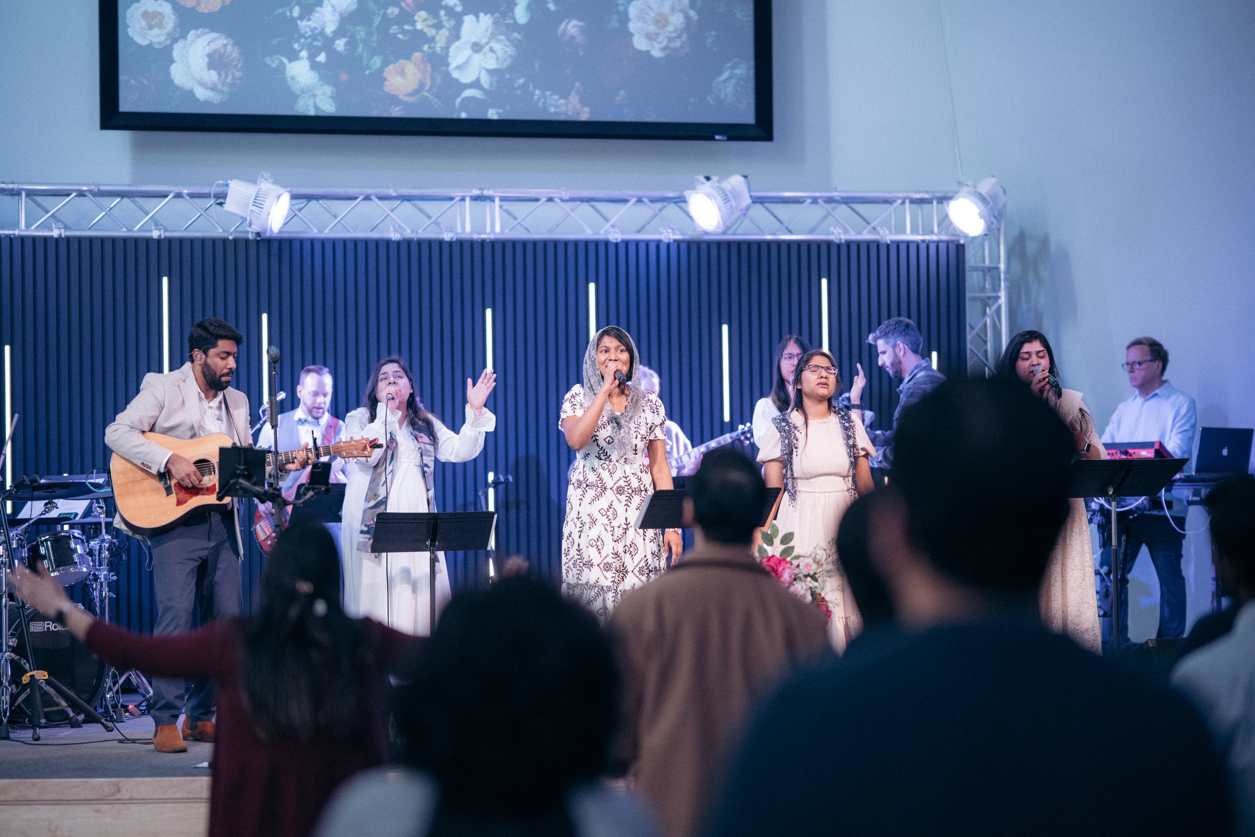 A group of people performing on stage at a church or concert, including a guitarist, singers, and musicians with a large screen in the background, and audience members visible in the foreground.