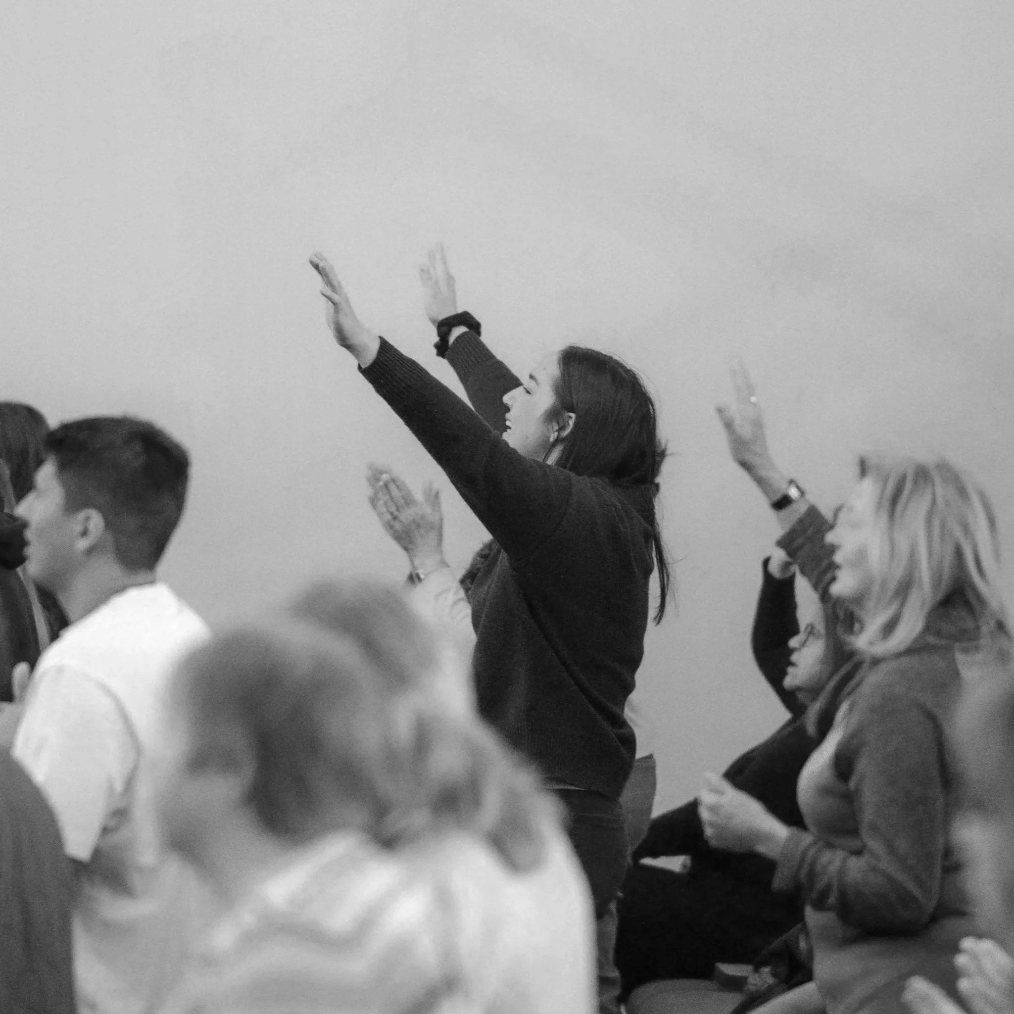 A woman with long hair and glasses raising her hand in a classroom or seminar setting, surrounded by others with similar gestures.