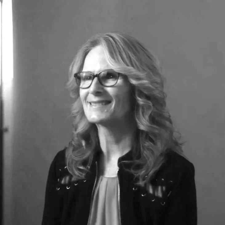 Black and white photo of a smiling woman with wavy hair and glasses.